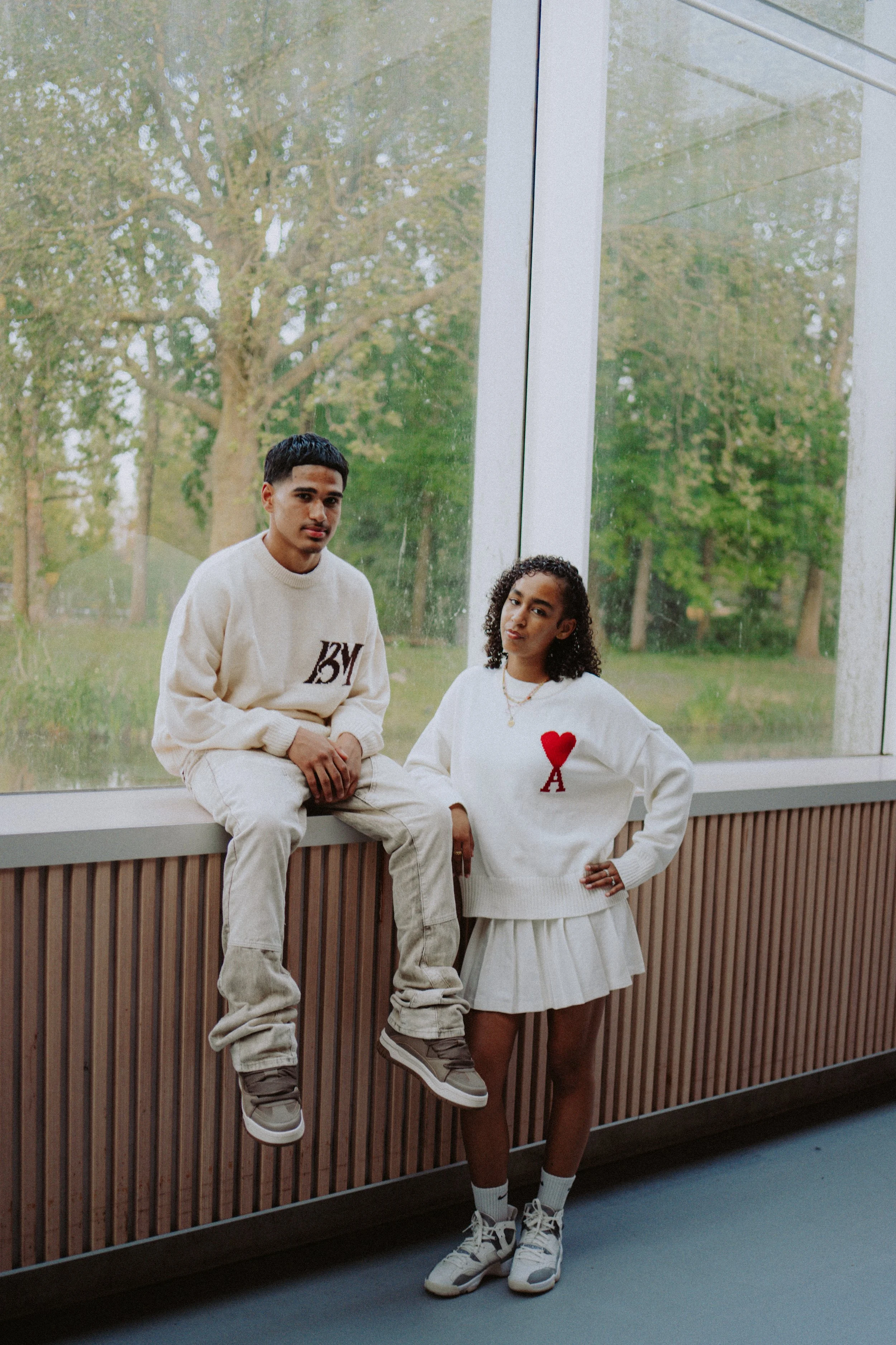 Two young people, a man and a woman, are indoors near large windows with a green outdoor scene in the background. The man is sitting on a ledge, wearing a cream-colored sweatshirt and pants, and sneakers. The woman is standing, wearing a white sweate