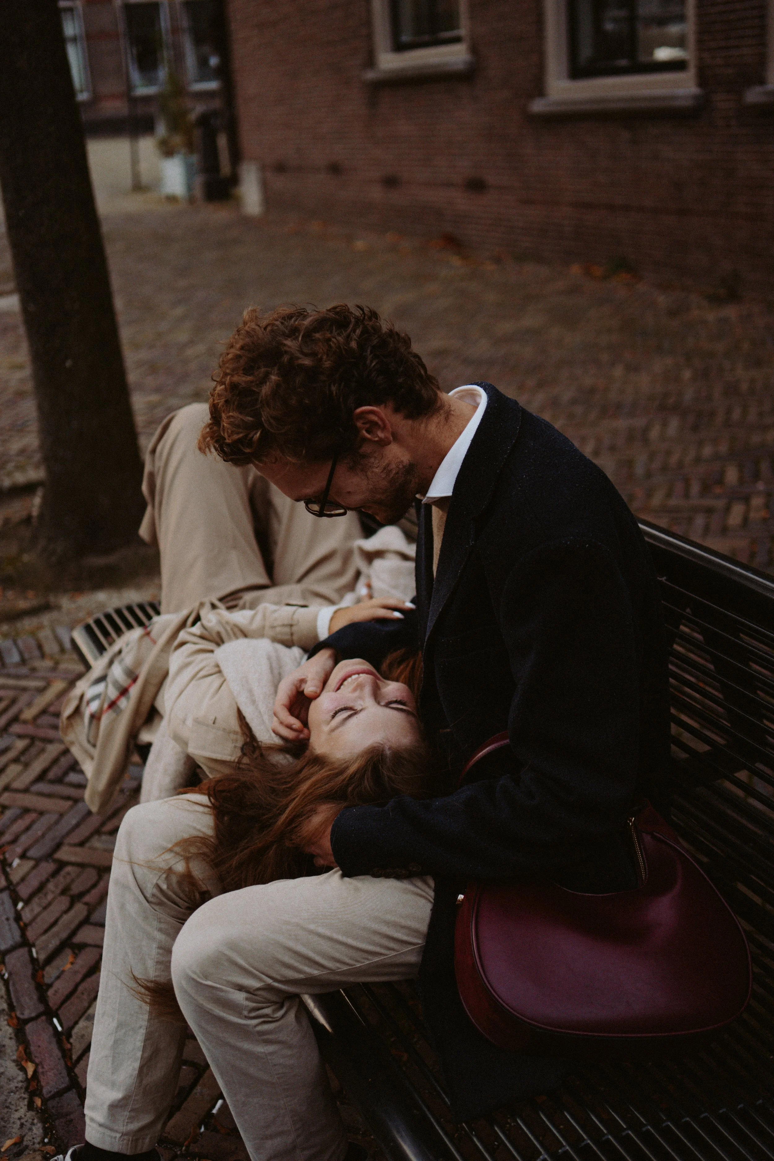 A couple is sitting on a park bench, leaning over each other with heads bowed, appearing to be comforting or sharing a moment of compassion. The man has curly hair and glasses, the woman has straight hair, and they are outdoors next to a brick buildi