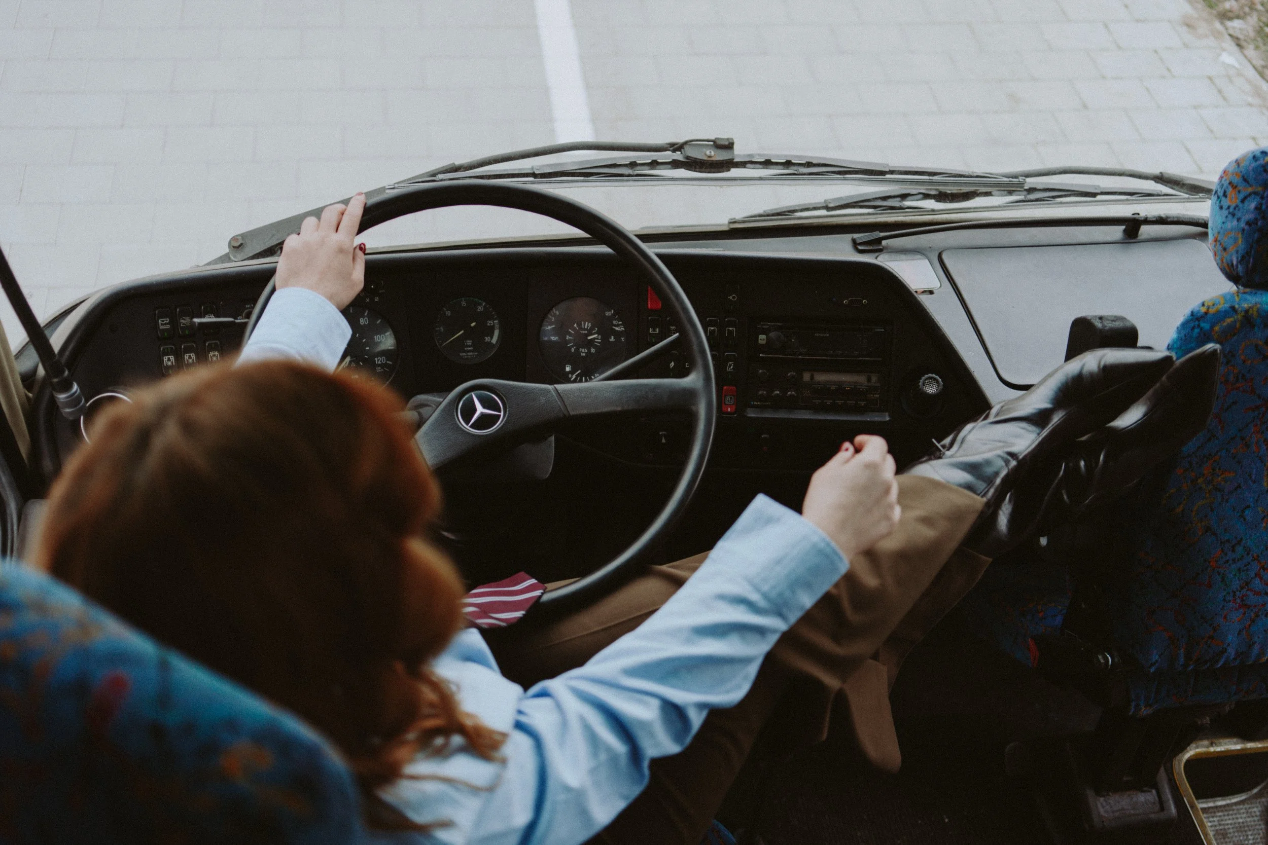 Interior of a bus or van showing dashboard and steering wheel with a Mercedes-Benz logo, a person's hand on the wheel, and another passenger in a patterned blue head covering.