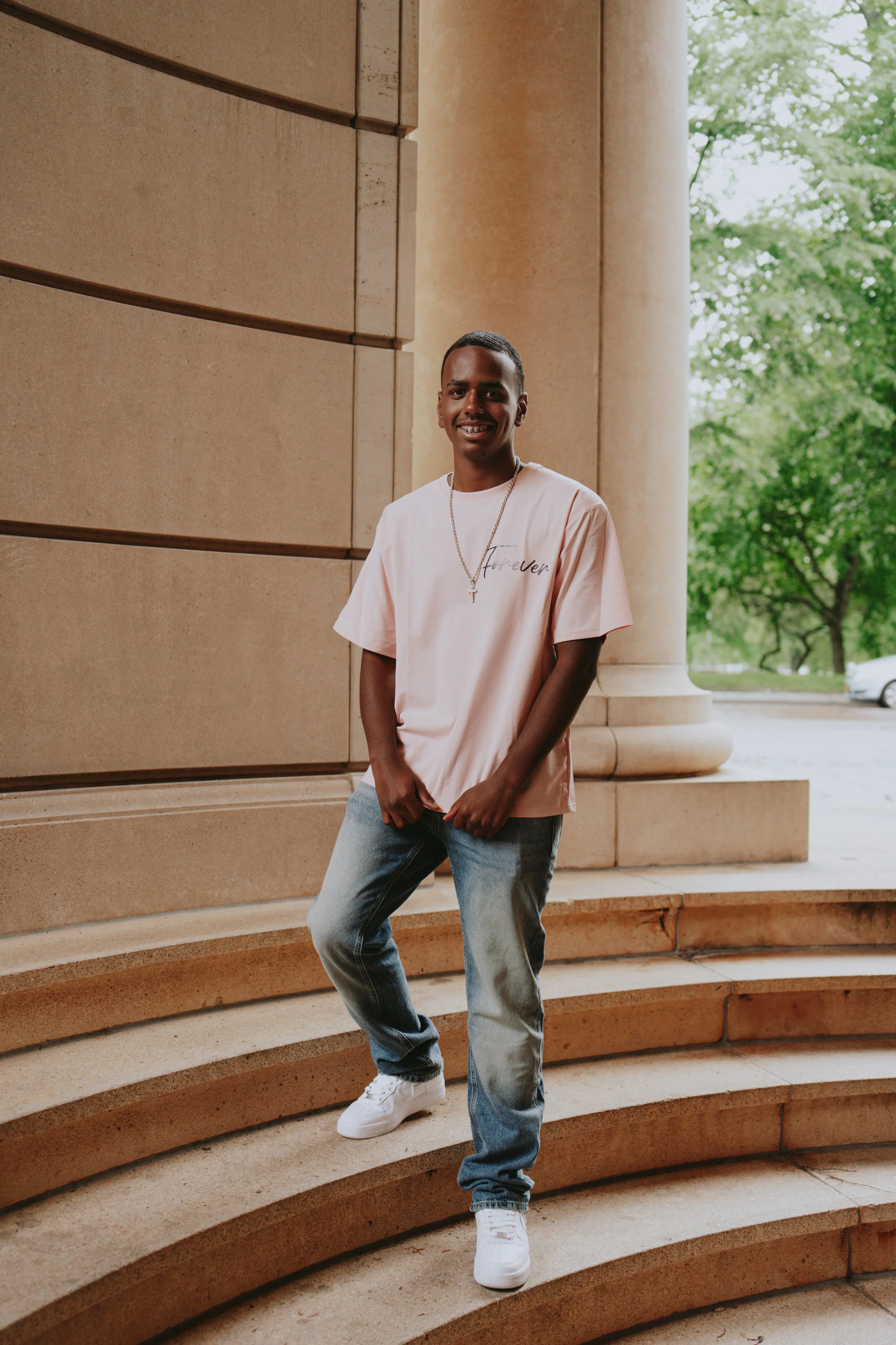 A young man standing on stone steps in front of a large building with tall columns and trees visible in the background. He is smiling and wearing a light pink T-shirt, jeans, white sneakers, and a necklace with a cross.