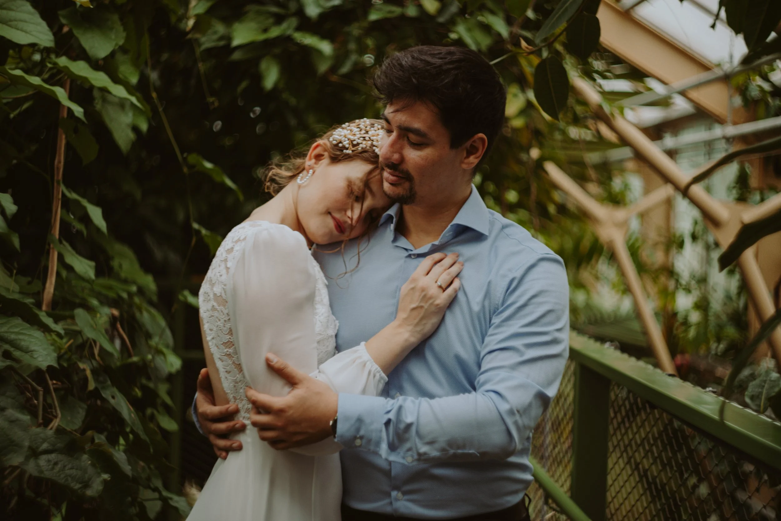 A man and woman sharing a tender moment in a green, plant-covered area, with the woman resting her head on the man's shoulder and both embracing.