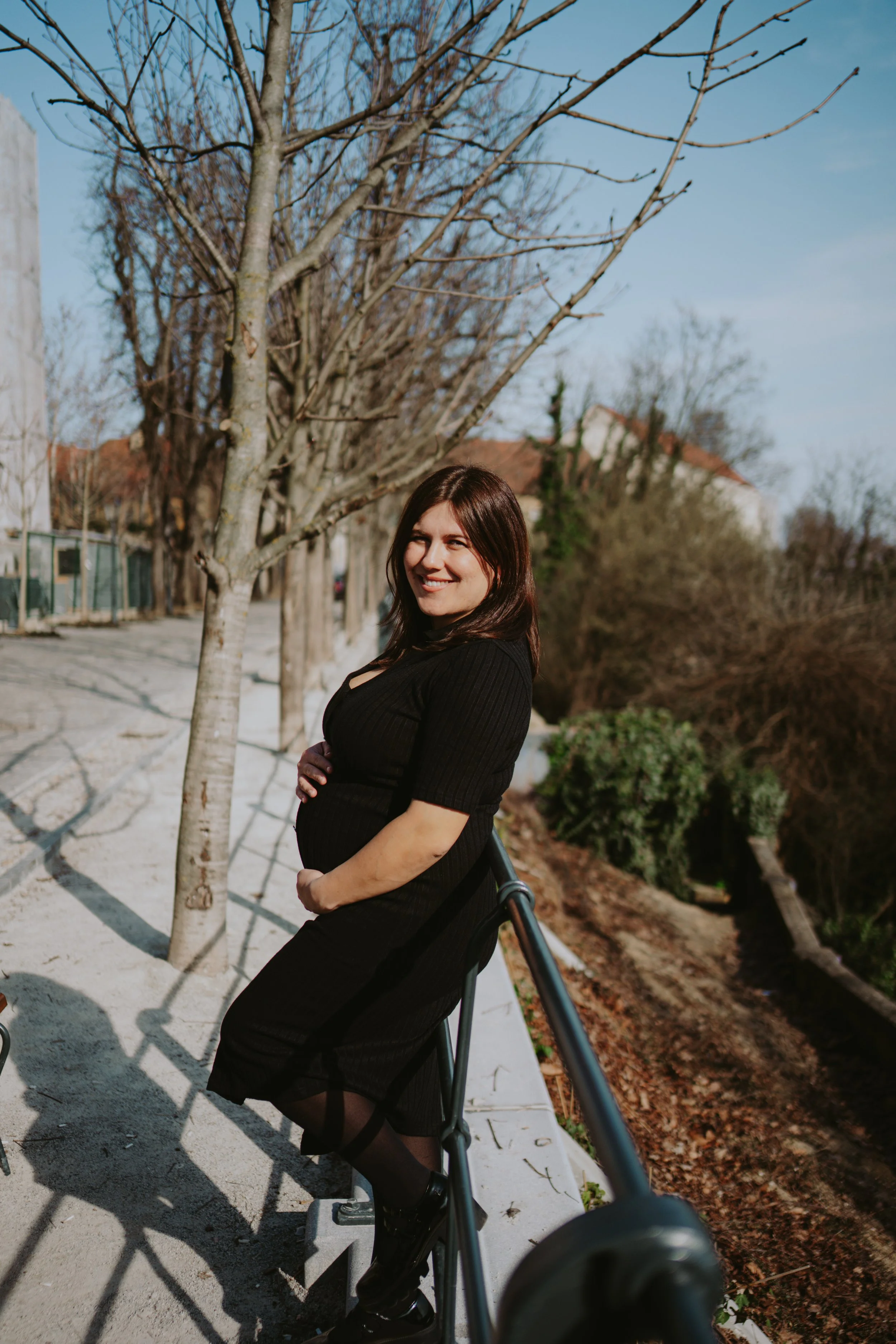 A pregnant woman smiling outdoors with trees and houses in the background, wearing a black dress and black shoes, leaning on a railing.