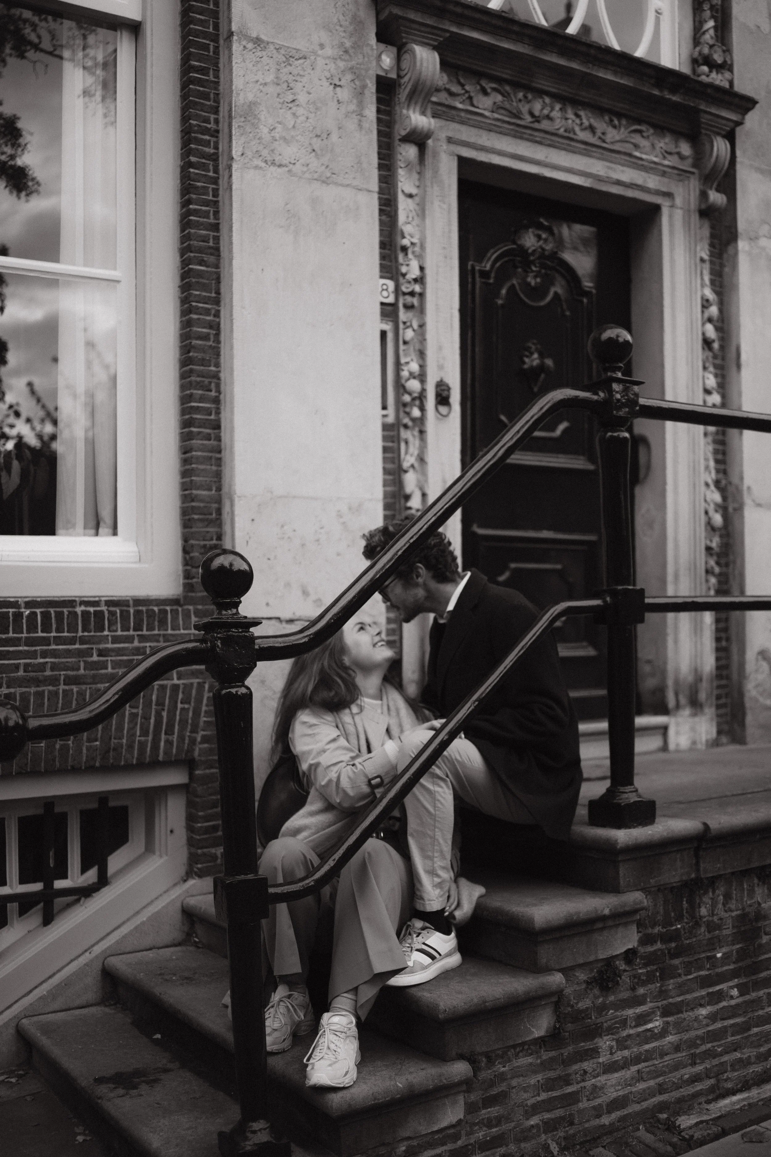 A couple sitting on stairs outside a building, looking at each other and smiling, with the man leaning towards the woman.