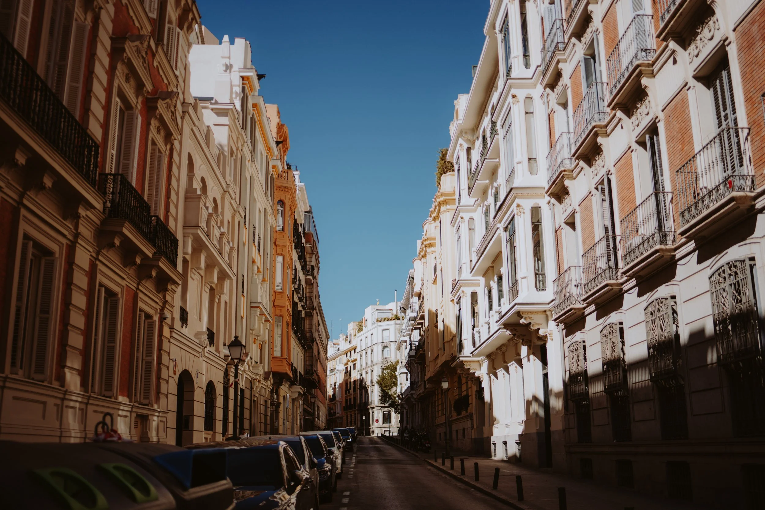 A narrow city street with parked cars on both sides, surrounded by tall, historic buildings with ornate facades and balconies, under a clear blue sky.