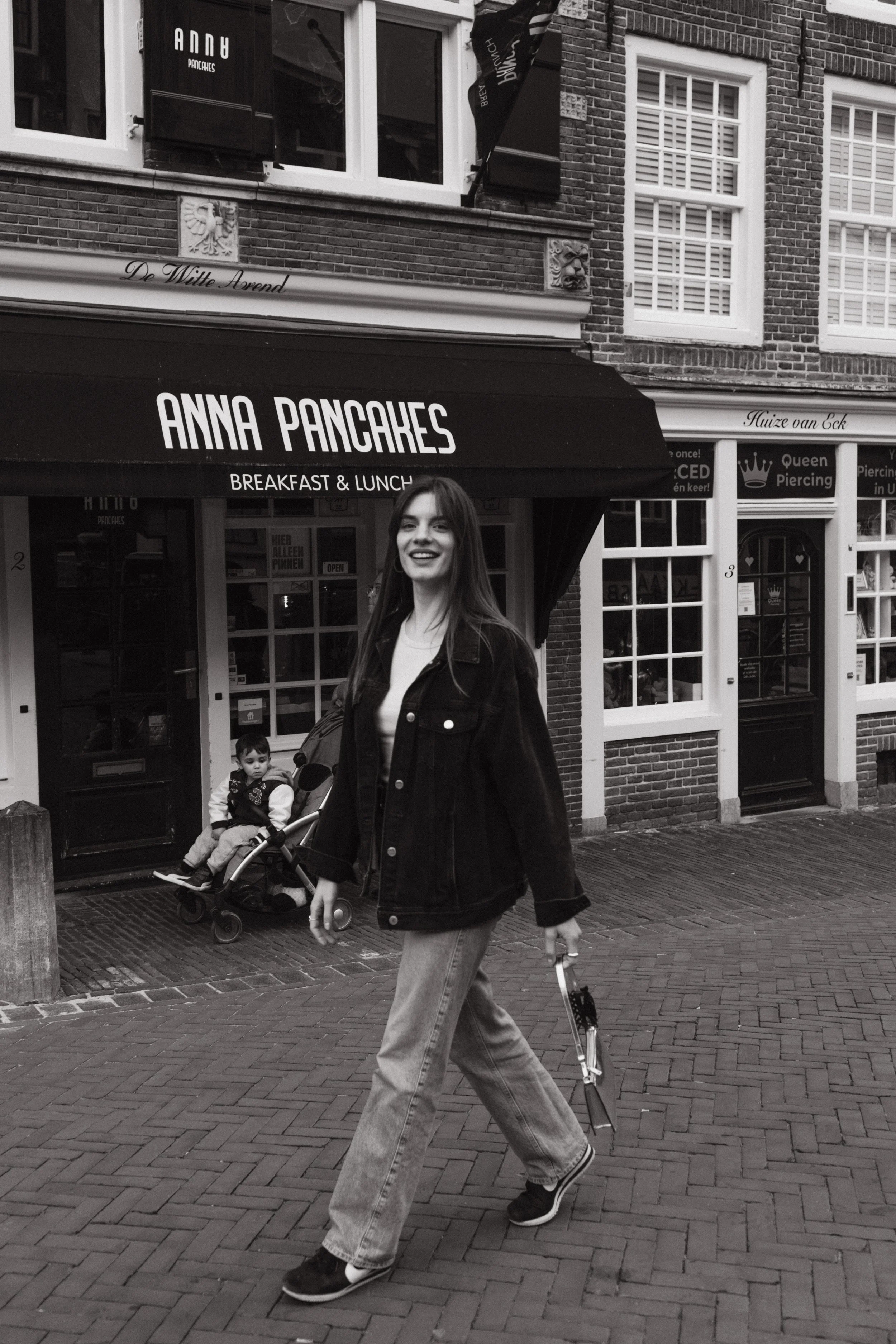 A woman walking across a city street while smiling at the camera. She is wearing a denim jacket, jeans, and sneakers. Behind her, there is a storefront with a sign that reads 'ANNA PANCAKES' and 'BREAKFAST & LUNCH'. There is also a child sitting in a