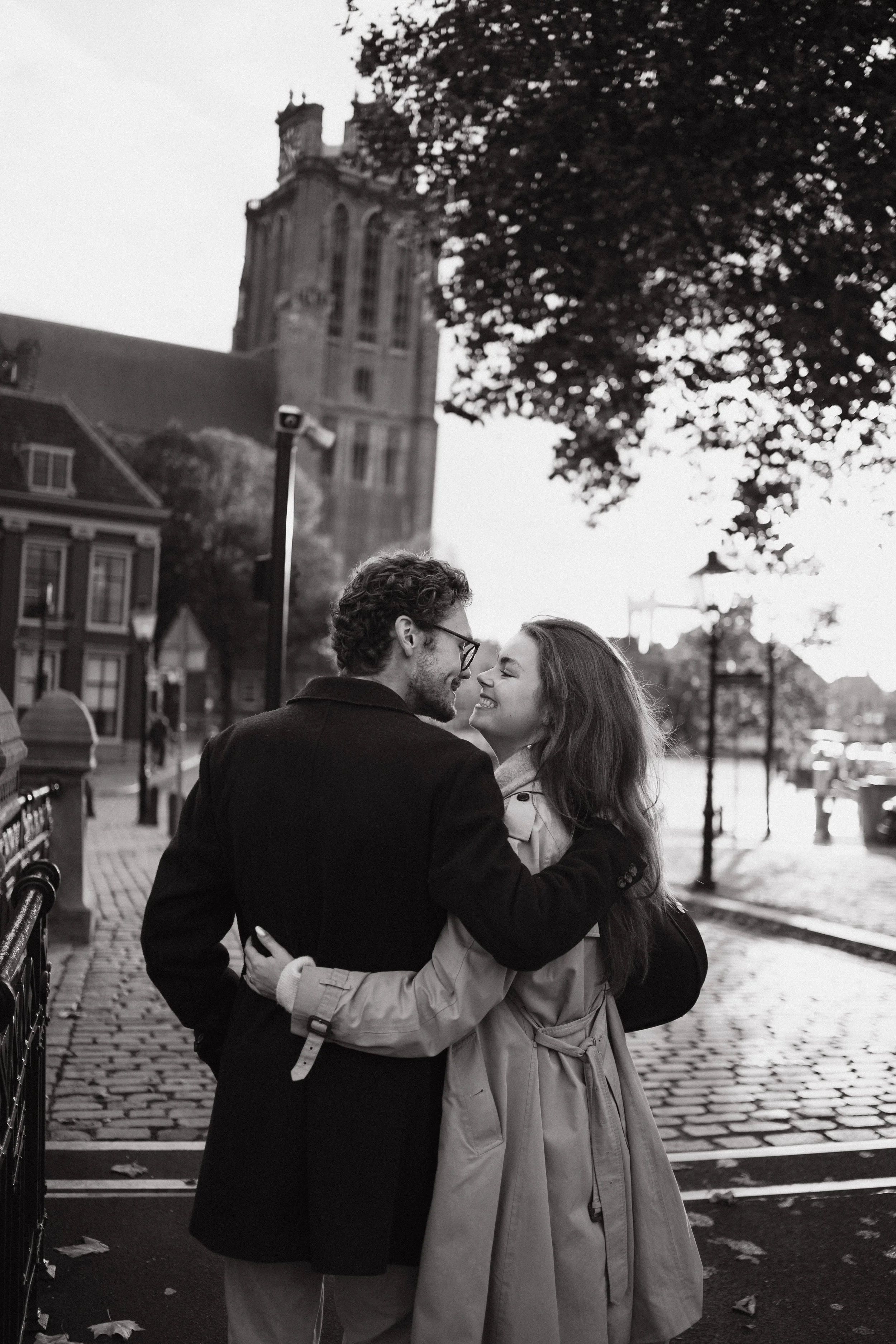 A black-and-white photo of a young couple smiling and embracing on a cobblestone street, with a historic church, trees, and cityscape in the background.