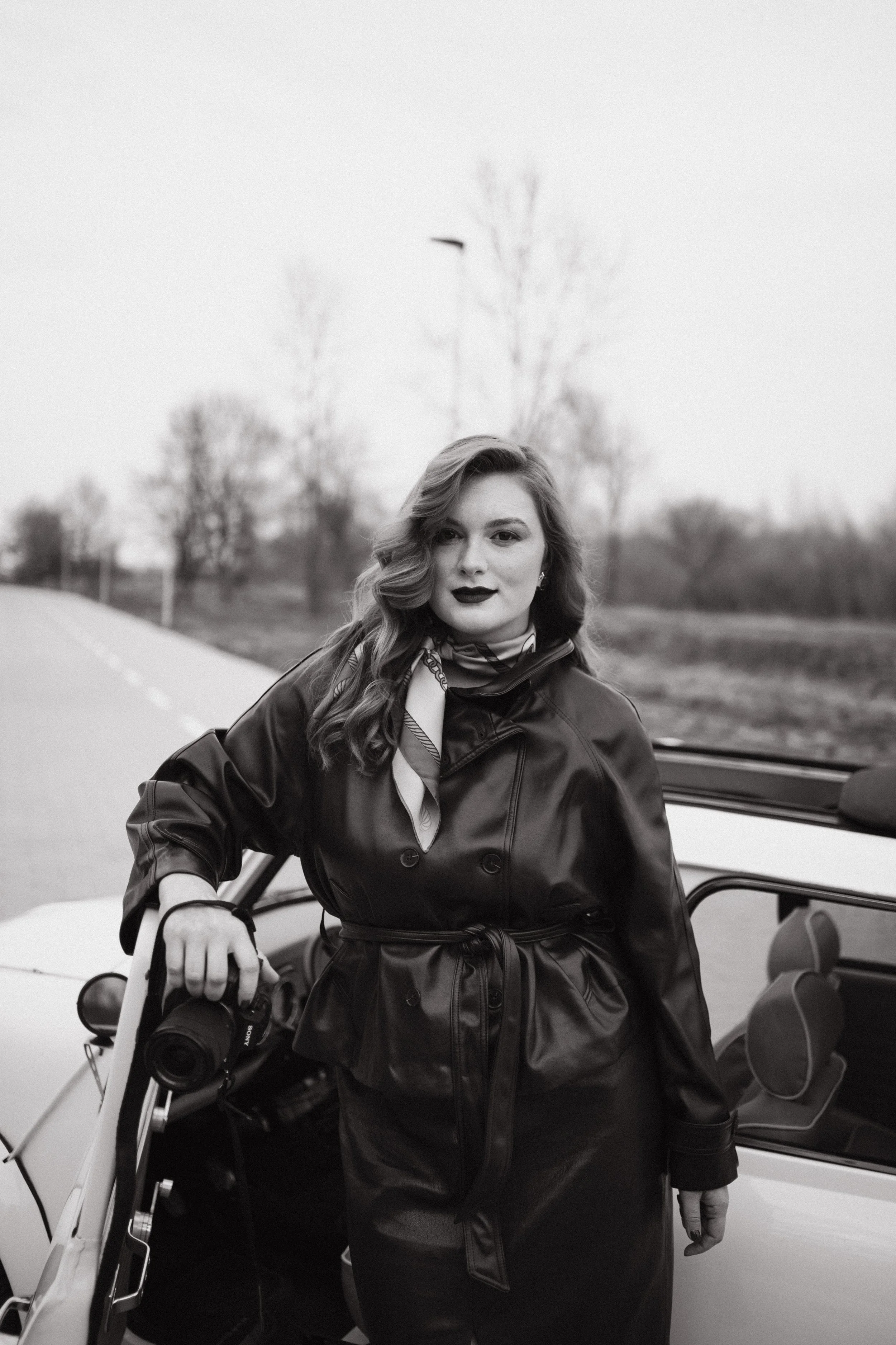 Branding photoshoot of a photographer. A woman with wavy hair wearing a leather coat, standing outside next to a vintage convertible car with doors open, holding a camera with a strap. 