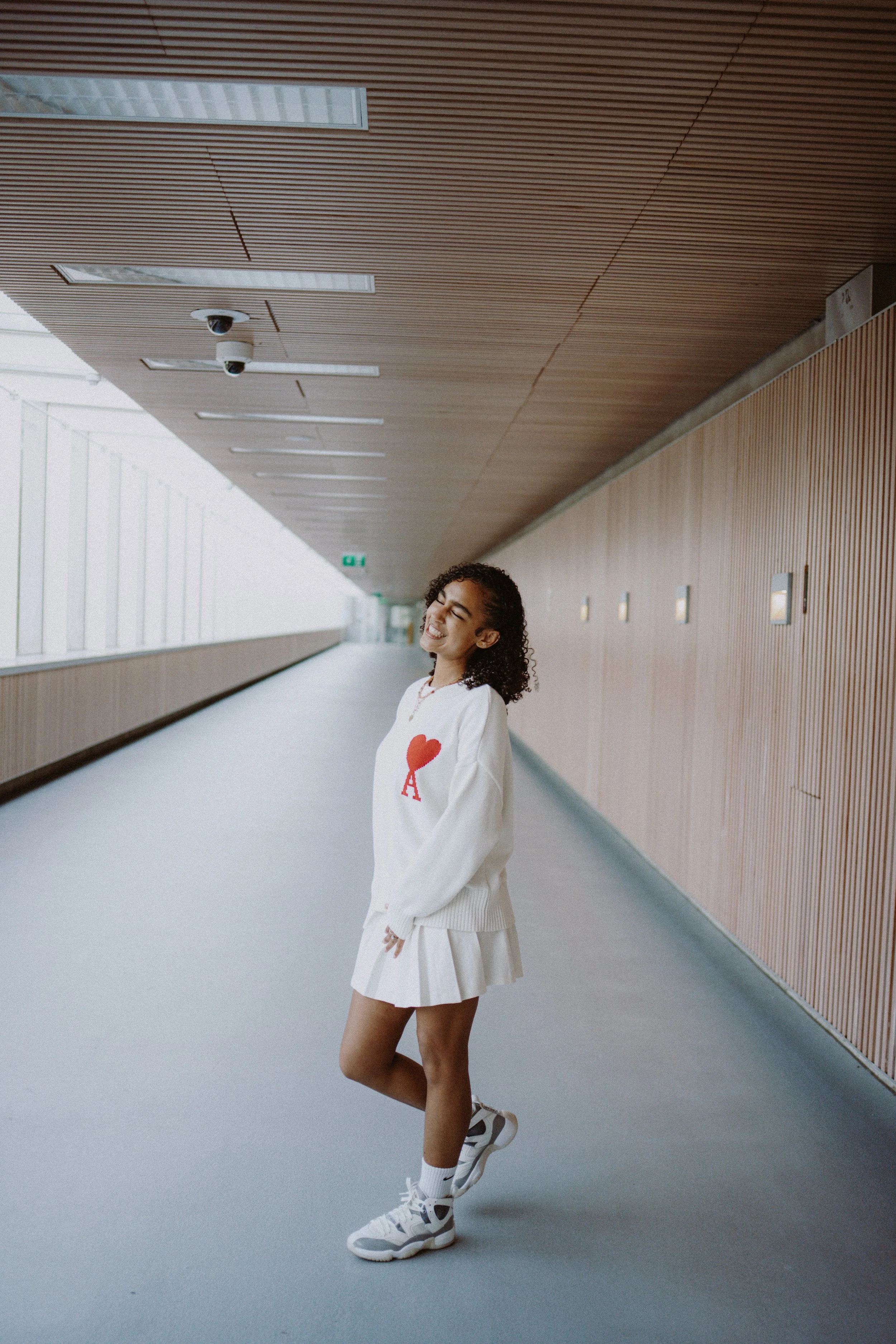 A young woman with curly hair smiling in a modern corridor, wearing a white sweatshirt with a red heart and letter 'A', a white skirt, white socks, and sneakers.