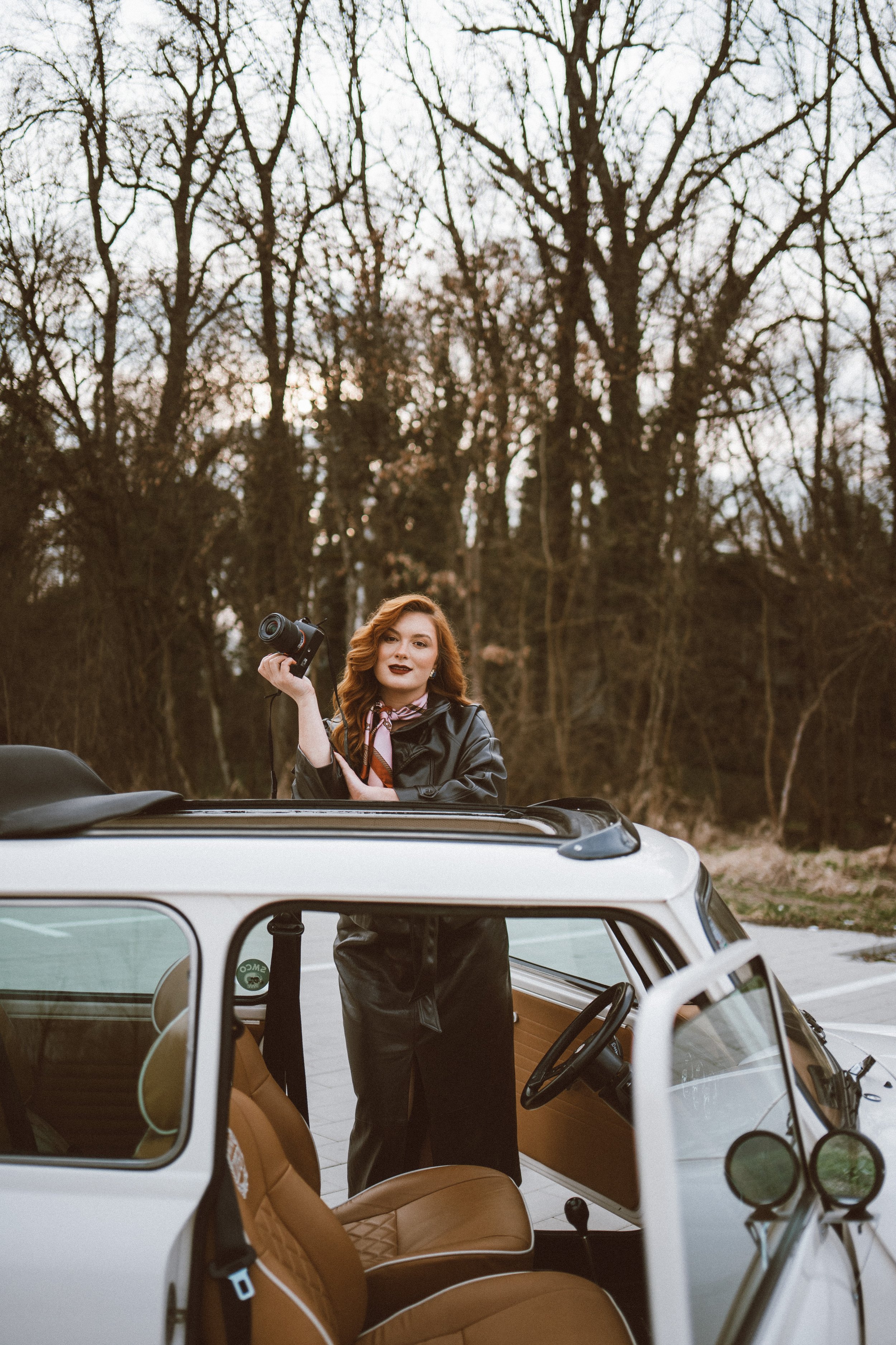 A woman with red hair and dark lipstick stands outside a white vintage car, holding a camera, in a natural setting with leafless trees in the background.