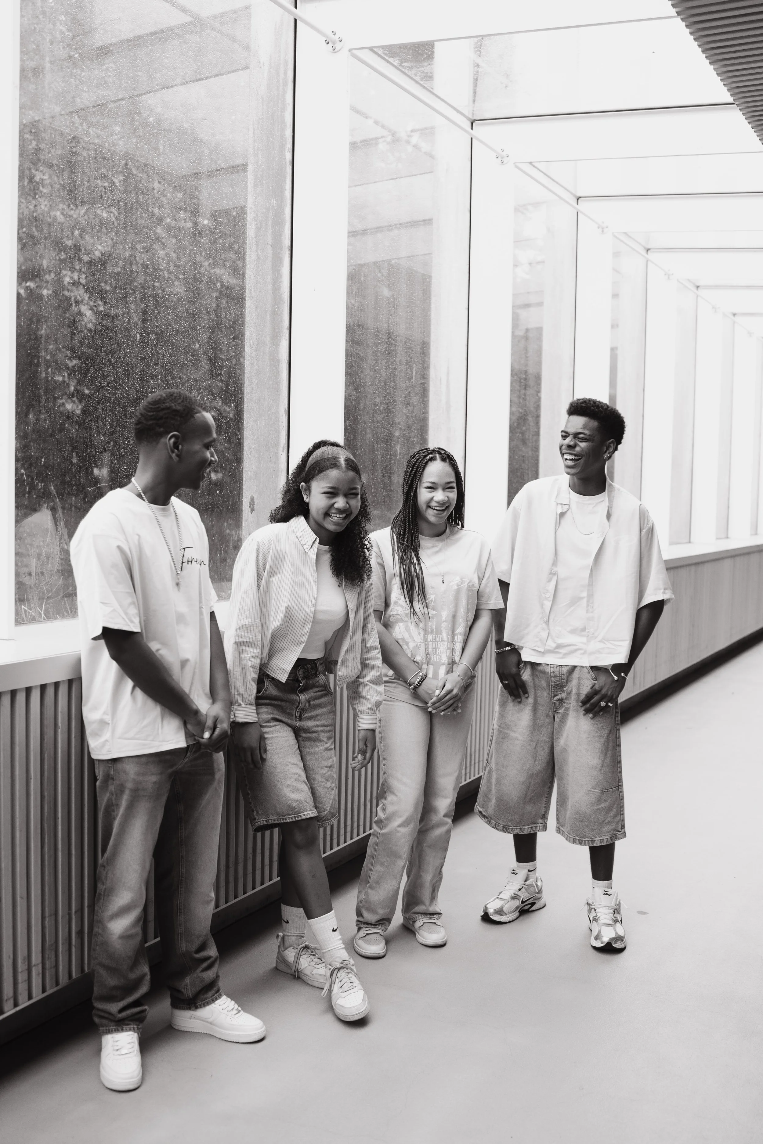Five young adults, three women and two men, standing in a modern hallway with large windows, smiling and laughing together.