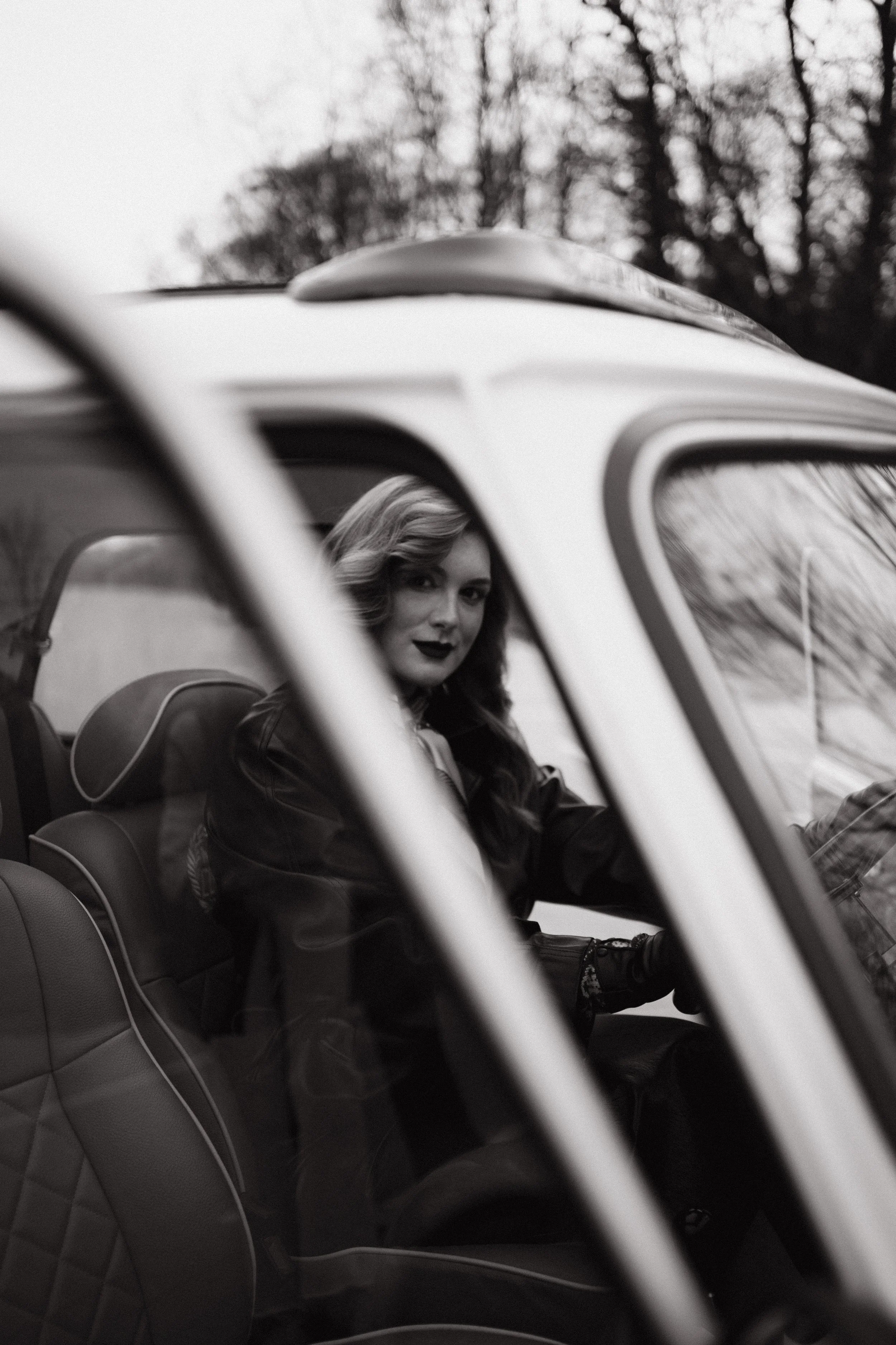 Black and white photo of a woman with wavy hair sitting in the driver's seat of a vintage convertible car, looking at the camera. Trees are visible in the background.
