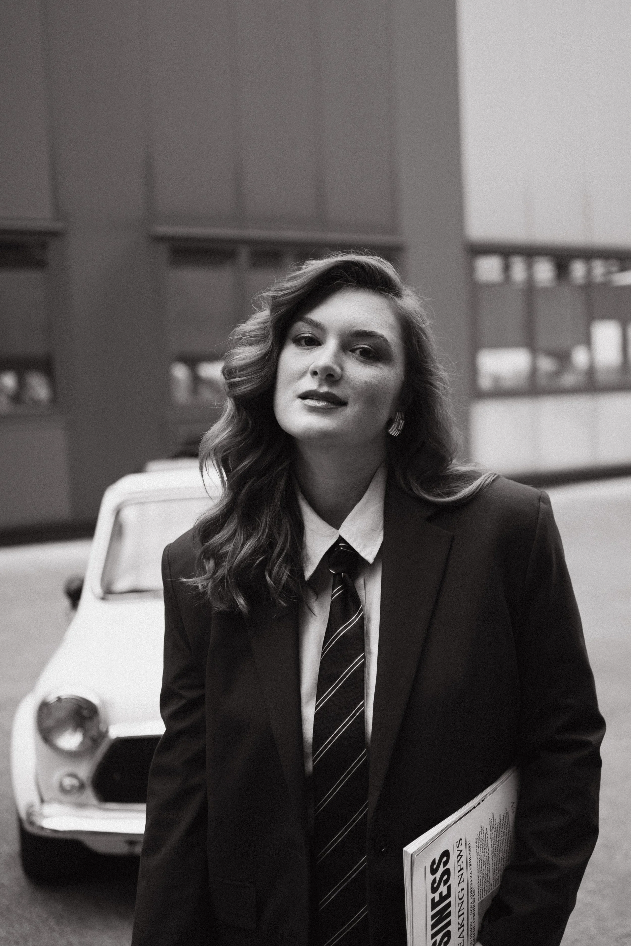 A woman in a business suit holding a magazine titled 'NEWS'. She is standing in front of a small vintage car in an indoor setting with large windows and modern architecture.