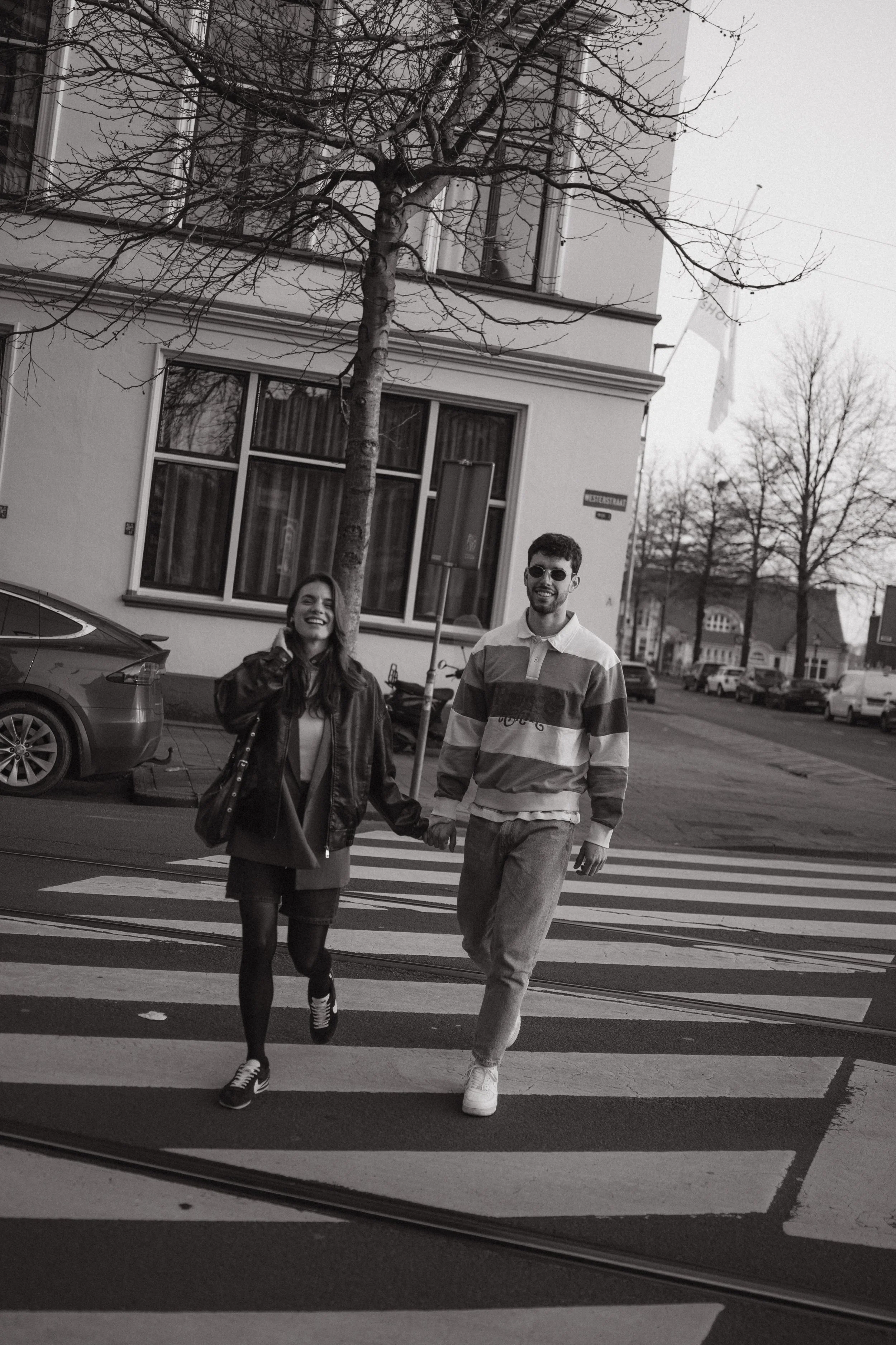 A black and white photo of a young couple holding hands and walking across a crosswalk on a city street, smiling and enjoying each other's company.