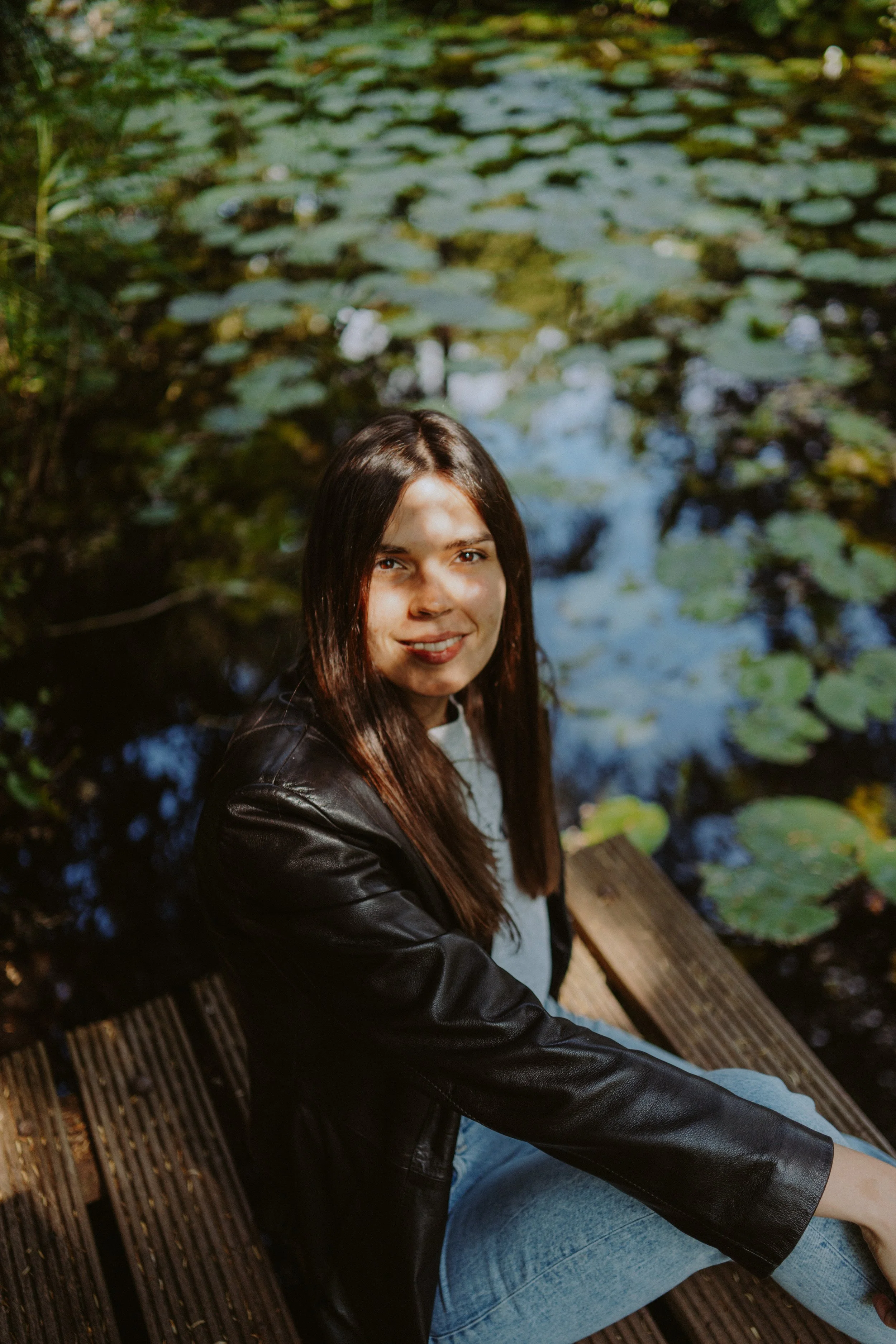 A young woman with long brown hair smiling while sitting on a wooden bench near water with lily pads.