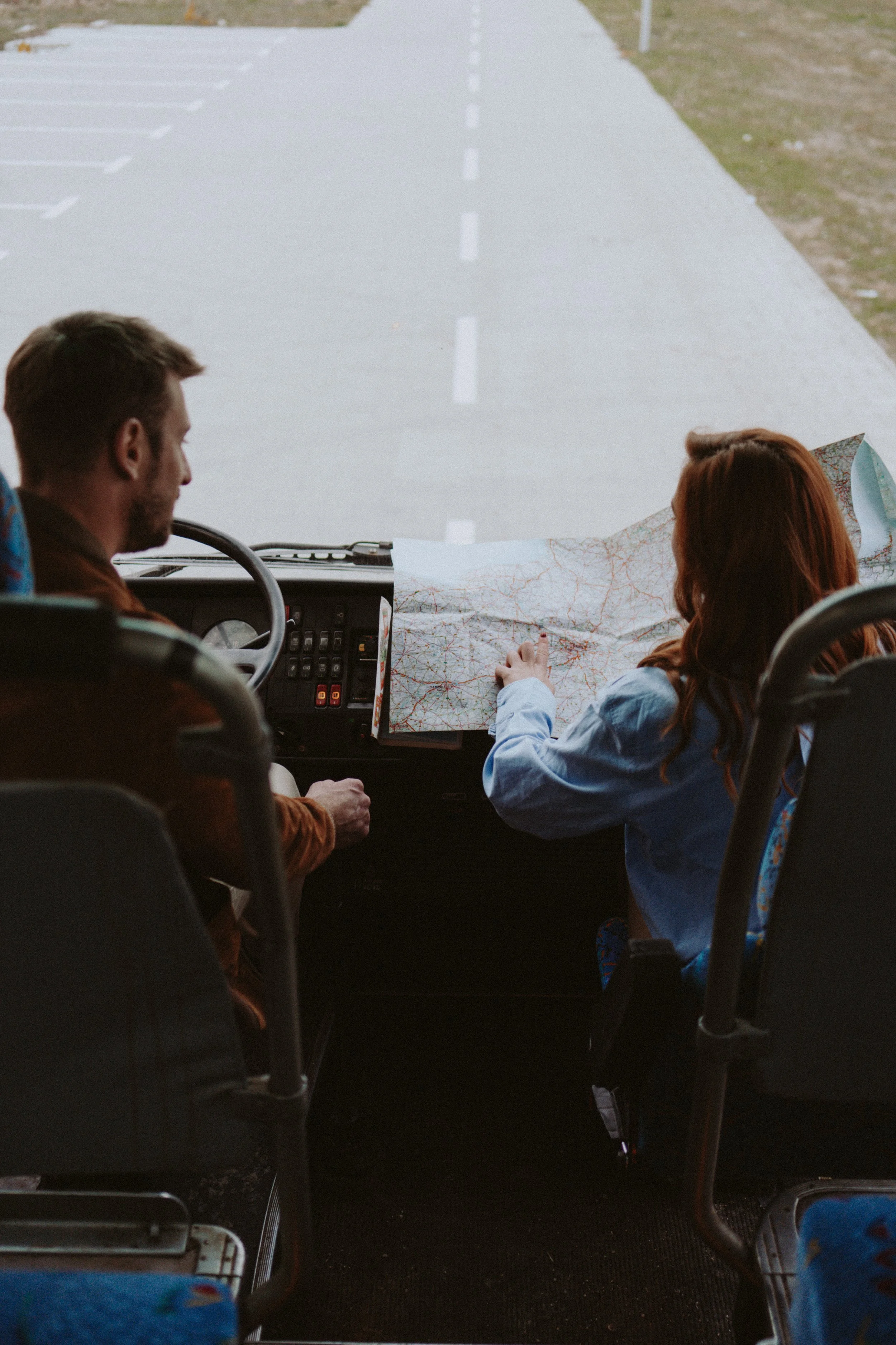 Two people sit in a bus looking at a map while driving on a road with marked lanes. The person on the left has short hair and a beard, and the person on the right has long red hair.