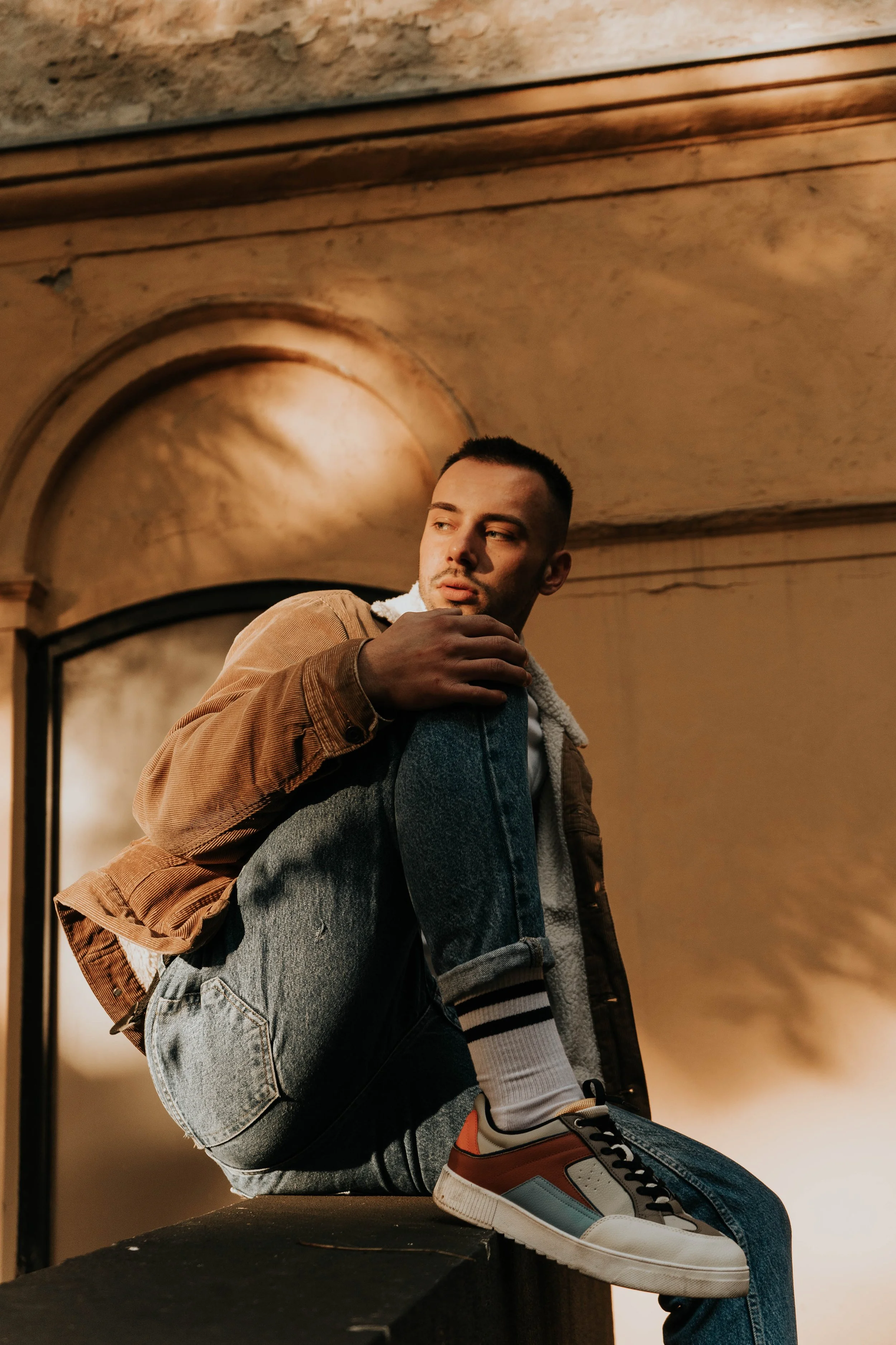 Young man with short dark hair, wearing a brown jacket, white hoodie, and sneakers, sitting on a black surface in a warm-toned room with ornate wall and ceiling details.