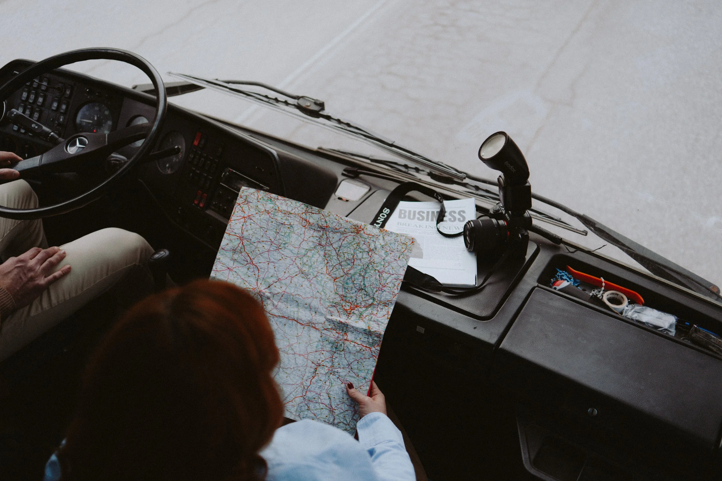 Interior of a bus with a driver and passengers, a route map, a newspaper, and a camera on the dashboard.