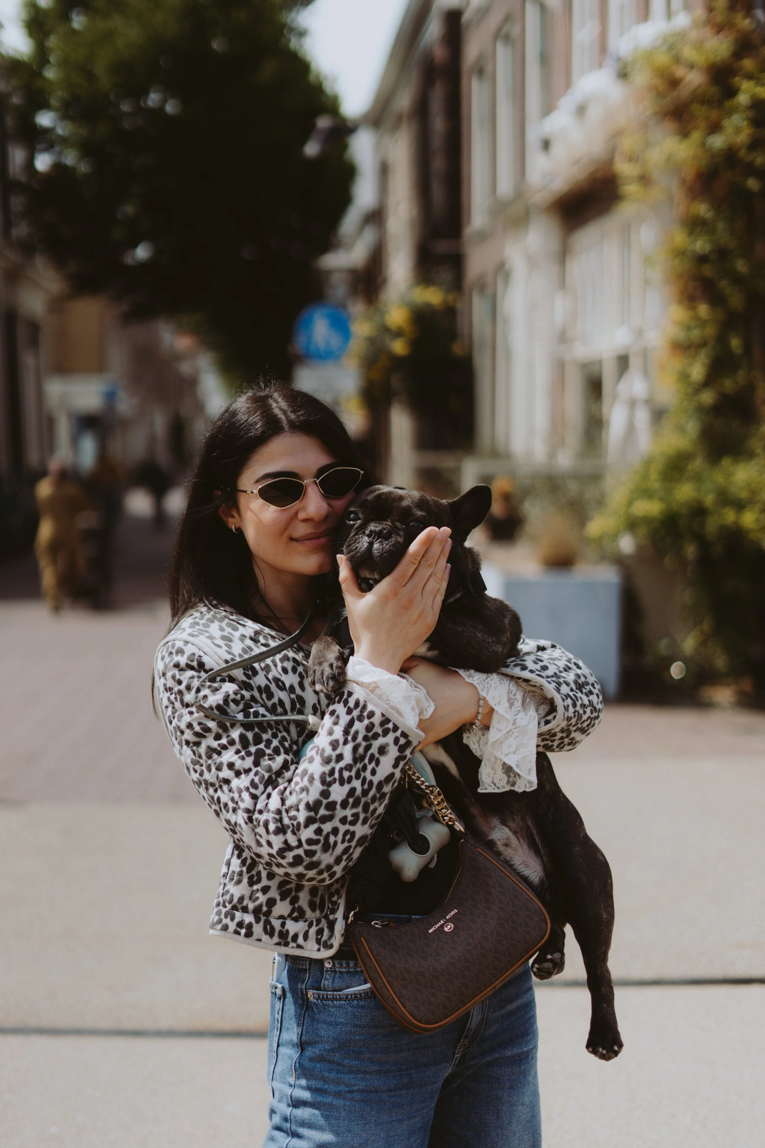 A woman with dark hair wearing sunglasses, a leopard-print jacket, and blue jeans, holding a small black French Bulldog in her arms.