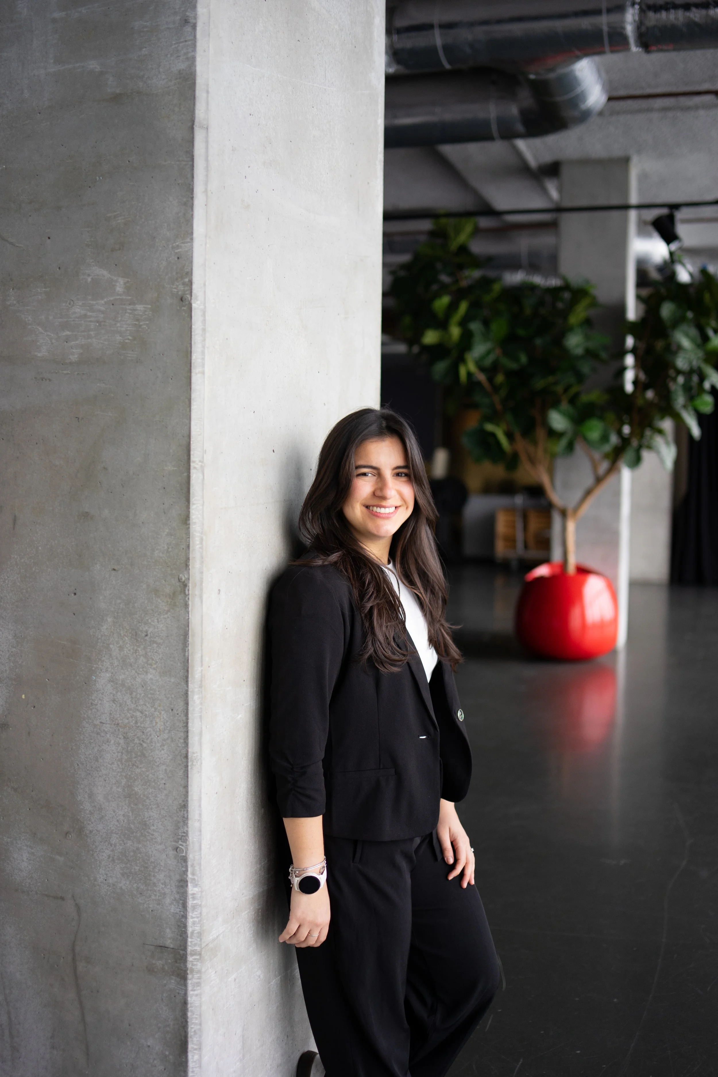 A smiling woman with long dark hair leaning against a concrete wall in a modern interior space with an industrial design, including exposed pipes and a large red potted plant in the background.