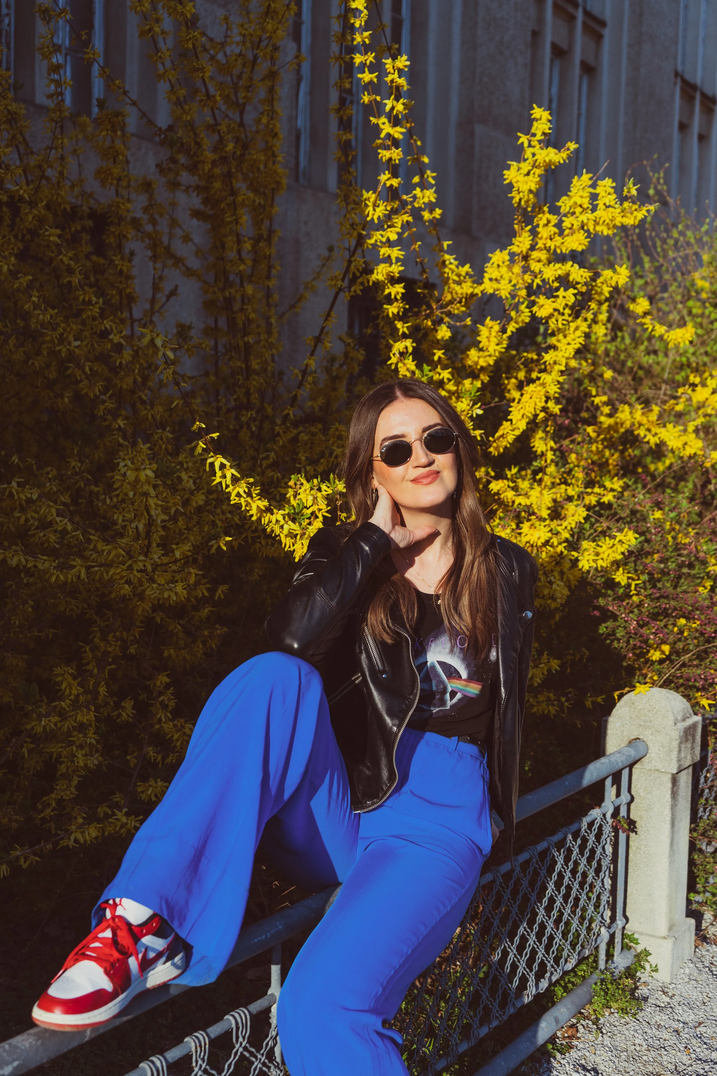 A woman sitting on a metal fence with her leg resting on it, outdoors at night. She is wearing sunglasses, a black leather jacket, a graphic t-shirt, bright blue pants, and red sneakers. There are yellow flowering bushes behind her and a building in 