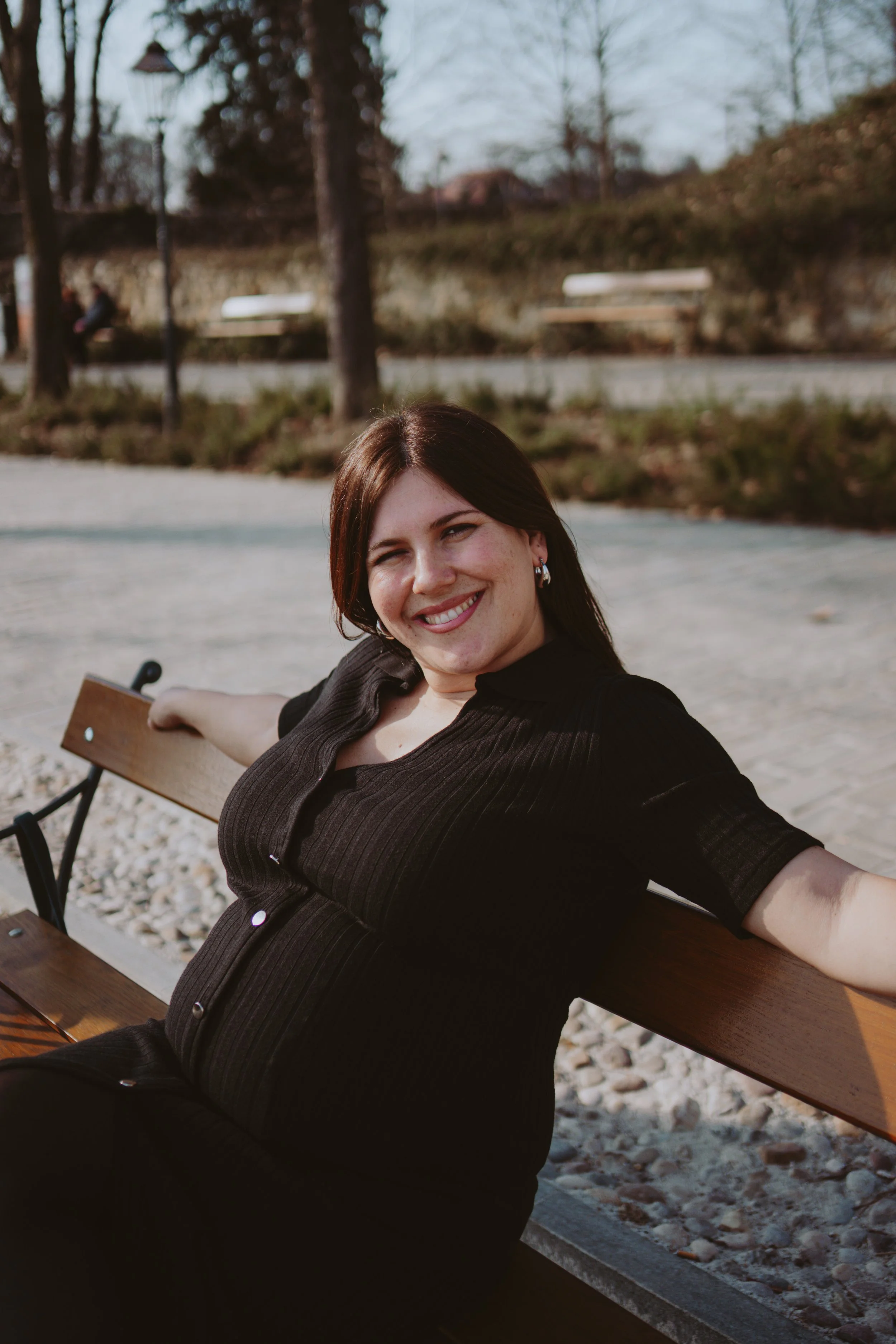 A woman with shoulder-length brown hair and hoop earrings smiling and sitting on a park bench during daytime. She is wearing a black top with buttons and is extending her arm across the back of the bench. In the background, there are trees, a person 