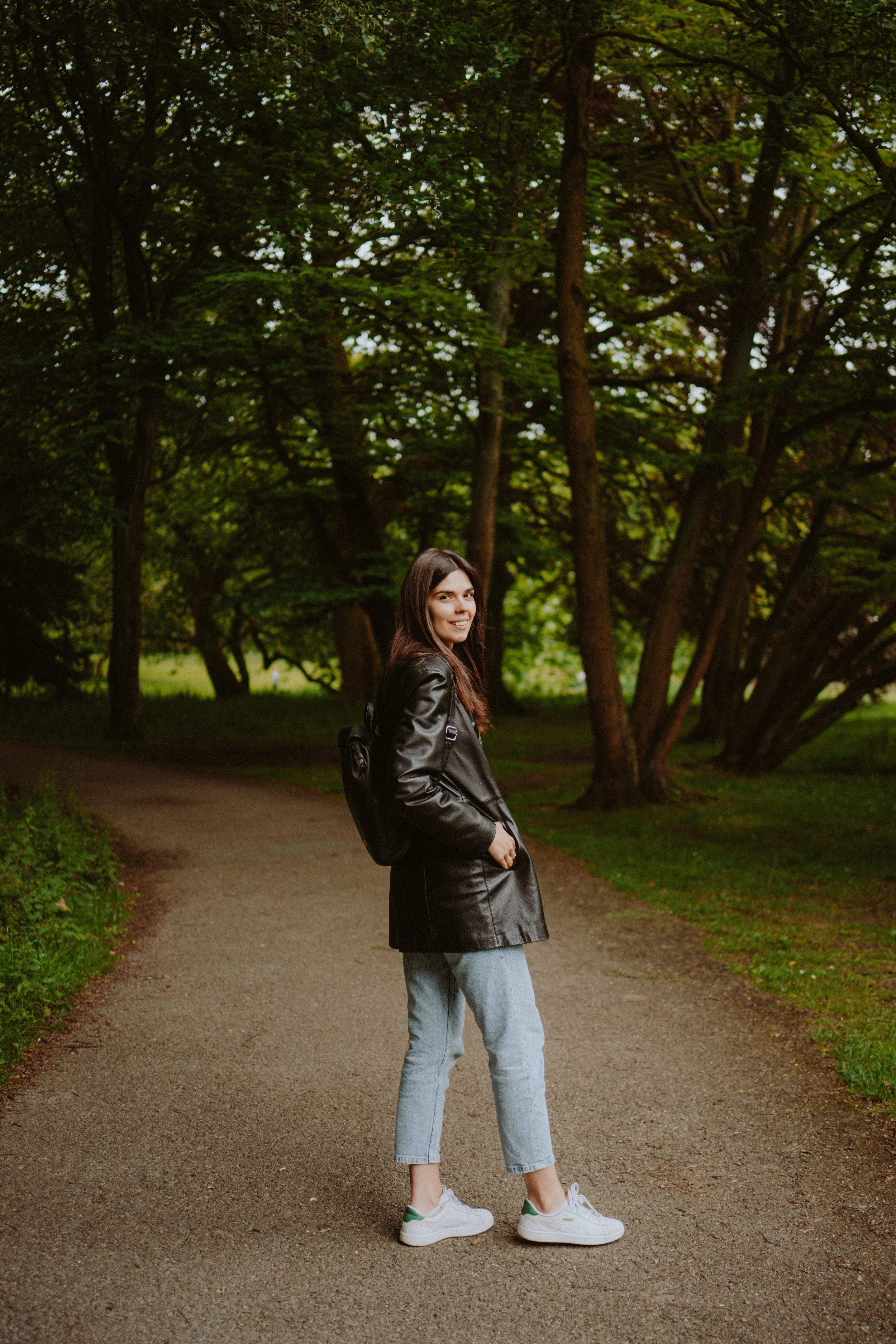 A young woman with long brown hair standing on a gravel path in a wooded park, smiling at the camera, wearing a black leather jacket, light blue jeans, and white sneakers with green accents, with her hands in her pockets.
