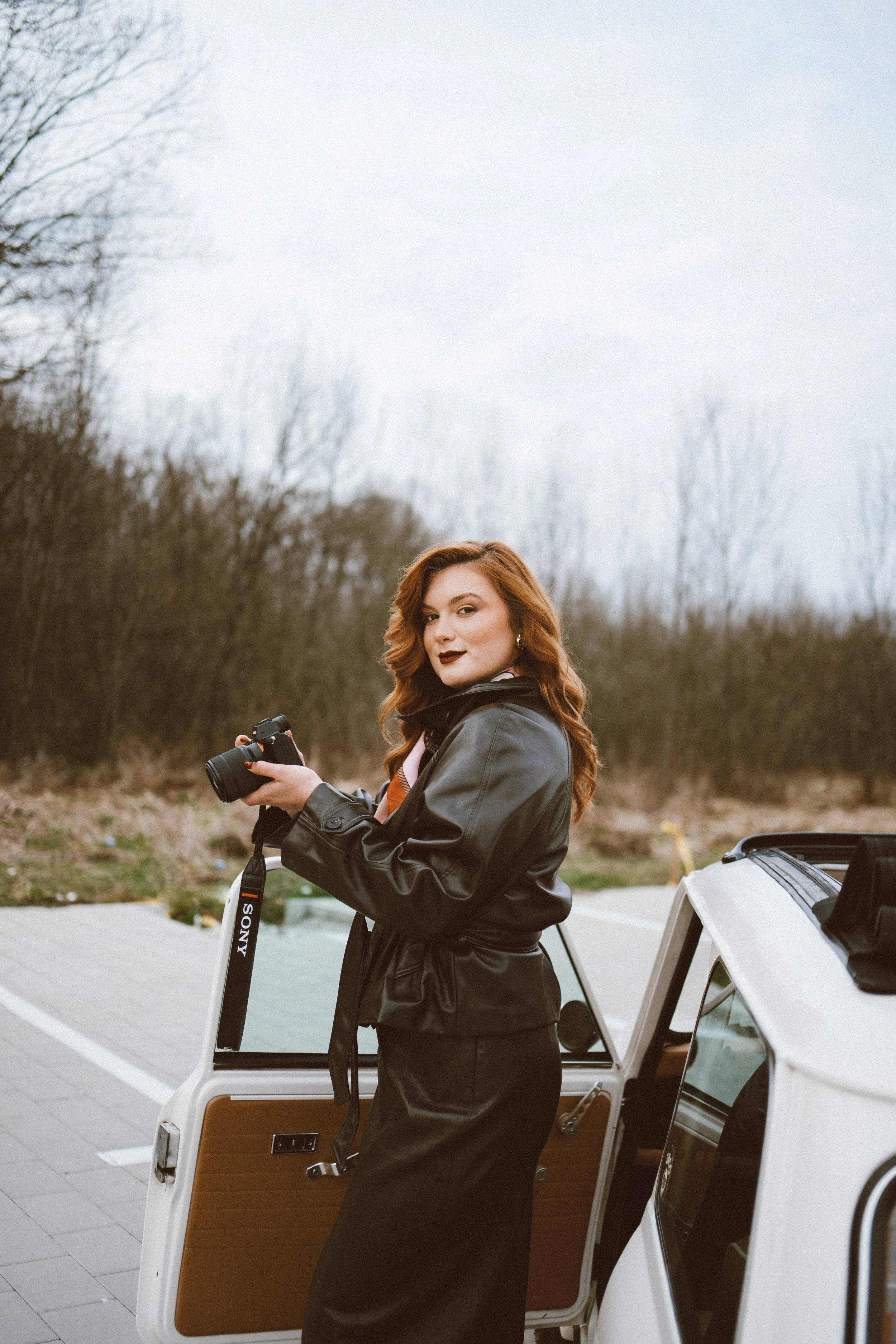 A woman with red hair holding a camera stands outside next to a white car with an open door. She is looking over her shoulder at the camera and is dressed in a black leather jacket and a long black skirt. The background shows leafless trees and an ov
