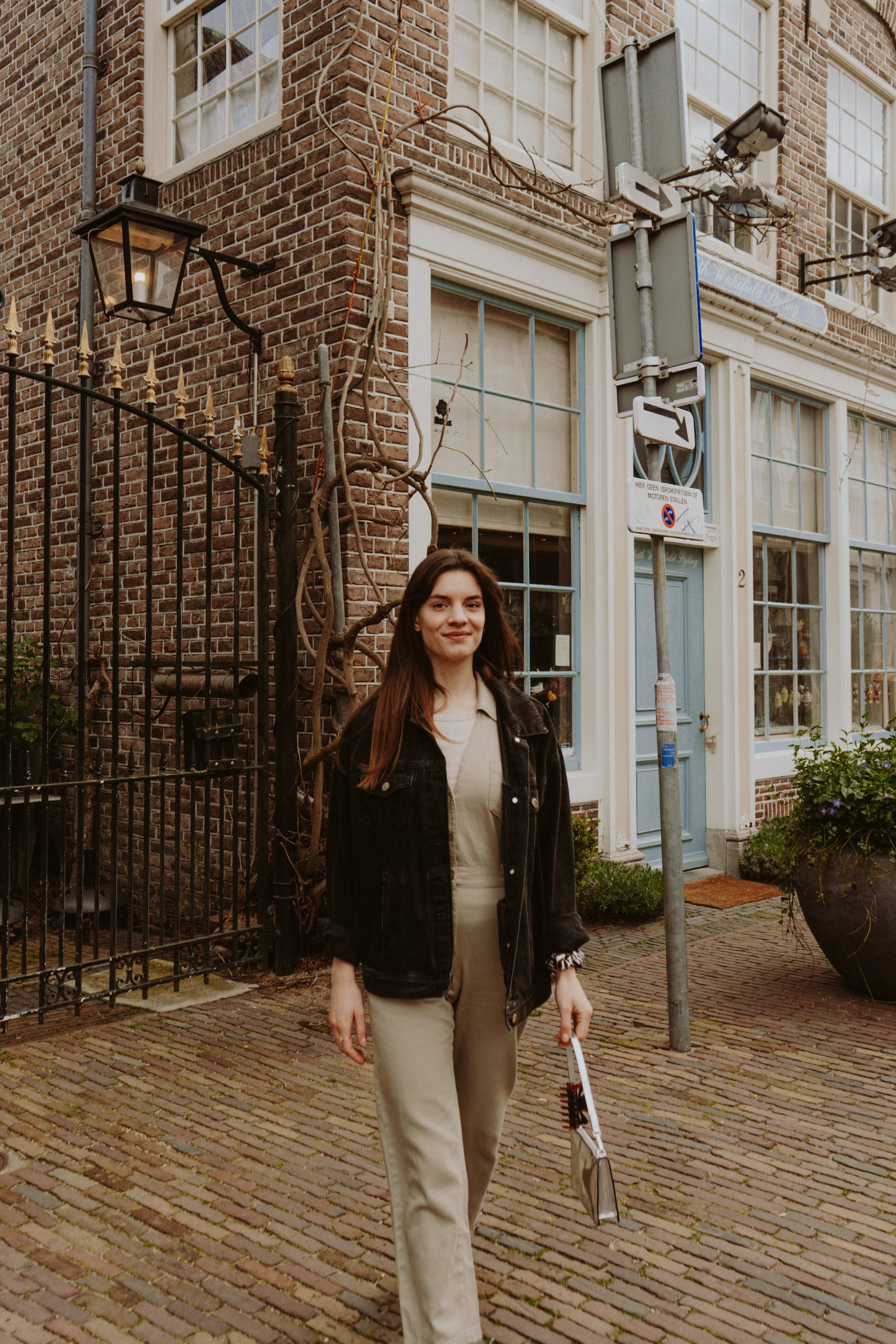 A young woman with long brown hair walking on a cobblestone street, carrying a white cane with a black tip, in front of a brick building with large windows and a blue door. There are street signs and a lamp post nearby.