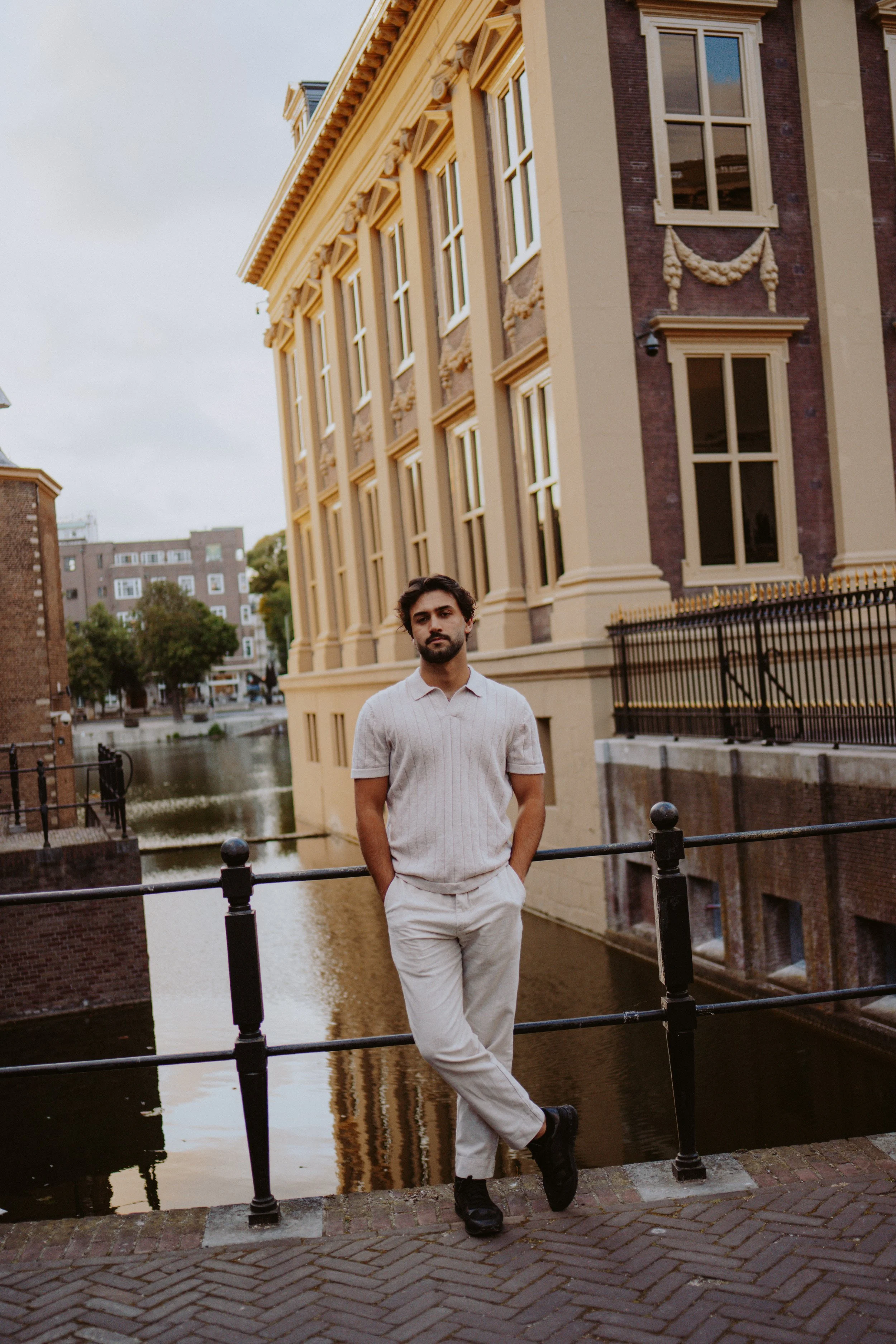 A young man with dark hair and beard, wearing a light-colored short-sleeve shirt and light pants, stands by a canal with his hands in his pockets. He is in front of a decorative building with beige trim and garland accents, which is beside a waterway