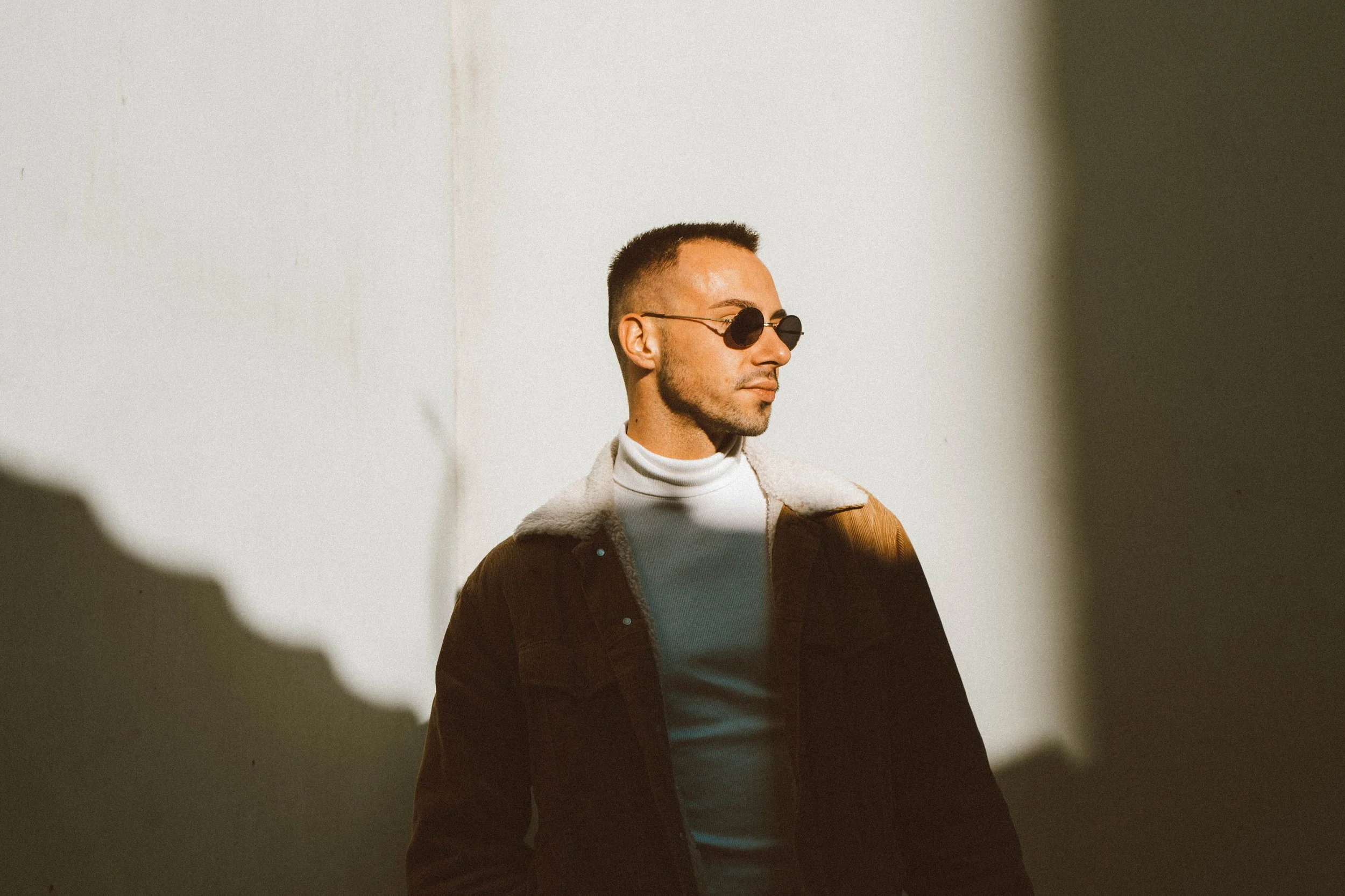 A man wearing round sunglasses and a white turtleneck under a brown jacket stands against a white wall, looking to the side with shadows cast on him from the lighting.