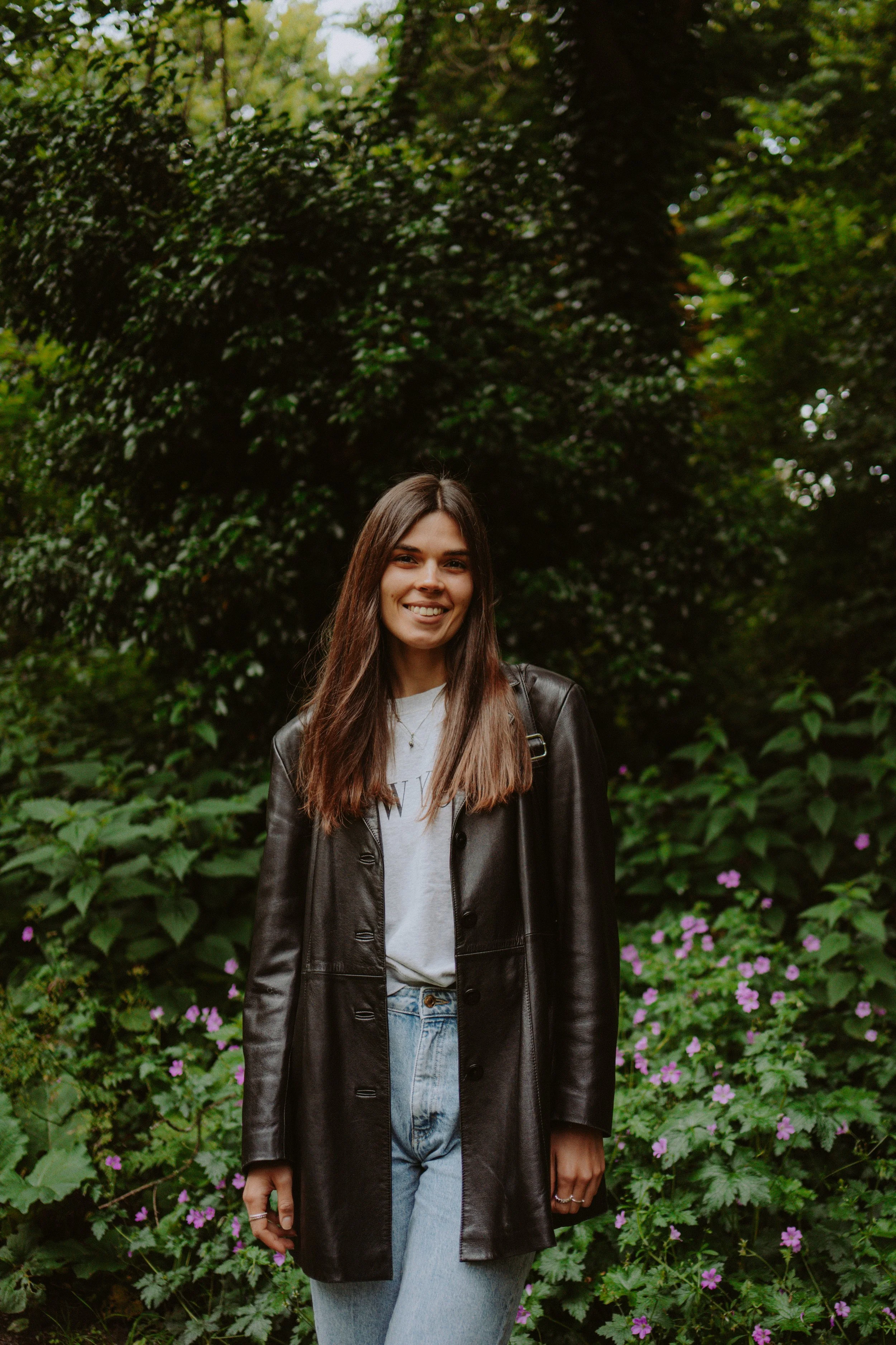 A young woman with long brown hair and fair skin standing outdoors in front of green foliage and purple flowers, smiling at the camera, wearing a leather jacket, white shirt, and light blue jeans.