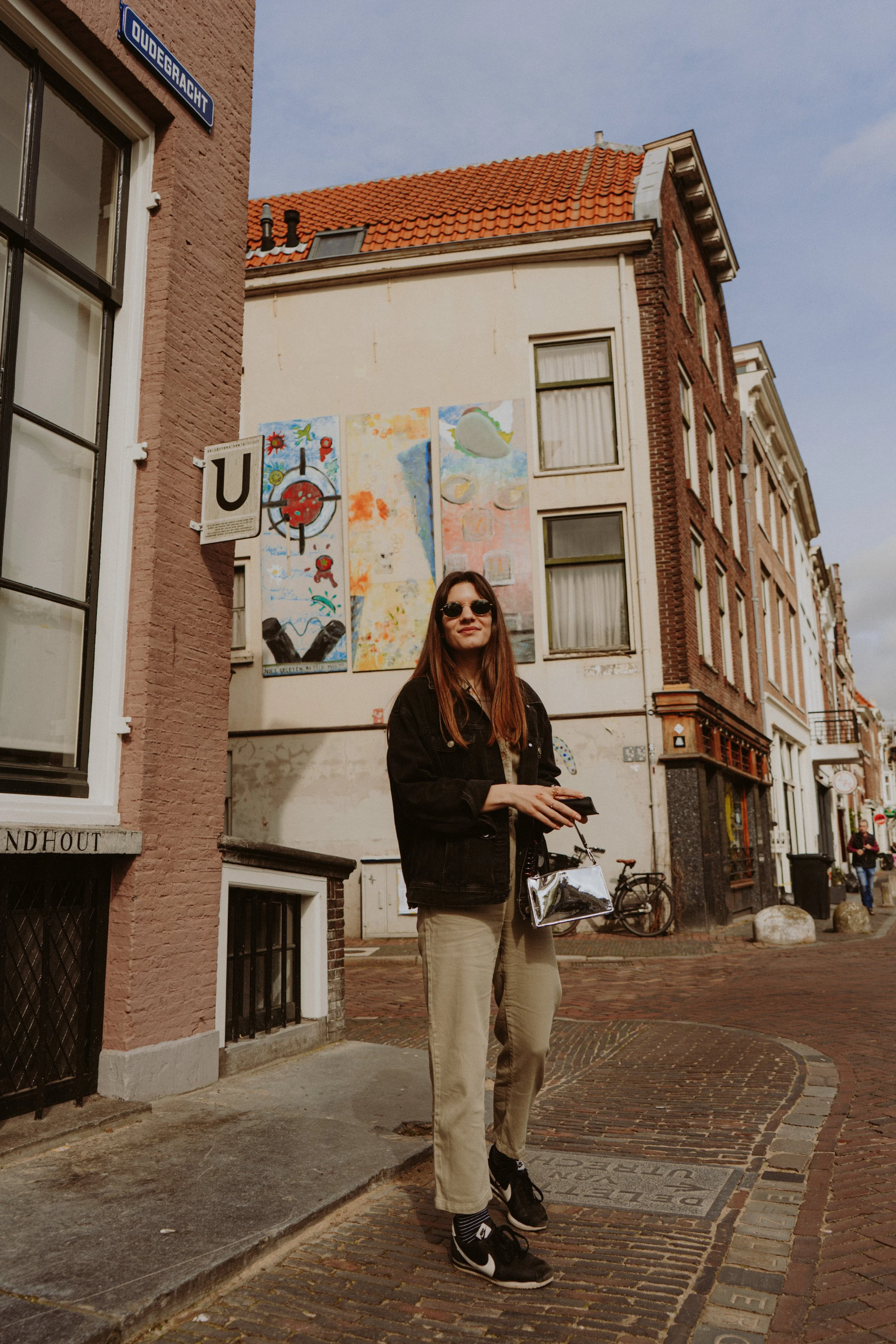 A woman wearing sunglasses, a black jacket, beige pants, and black sneakers standing on a cobblestone street in front of a mural on a building, with bicycles parked behind her.