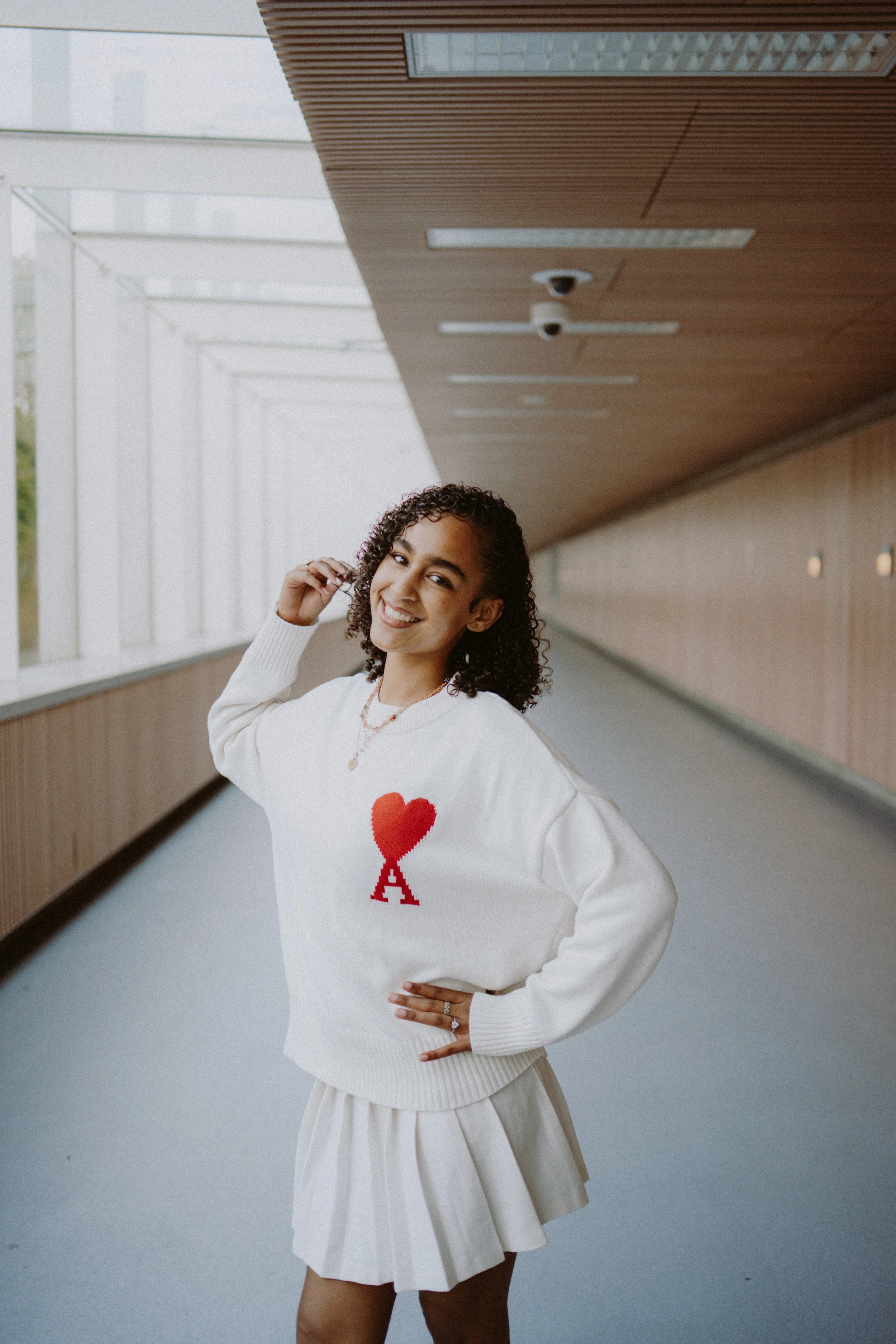 A young woman with curly hair smiling, wearing a white sweater with a red heart and letter A, and a light-colored skirt, standing in a modern corridor with wooden ceiling panels and large windows.