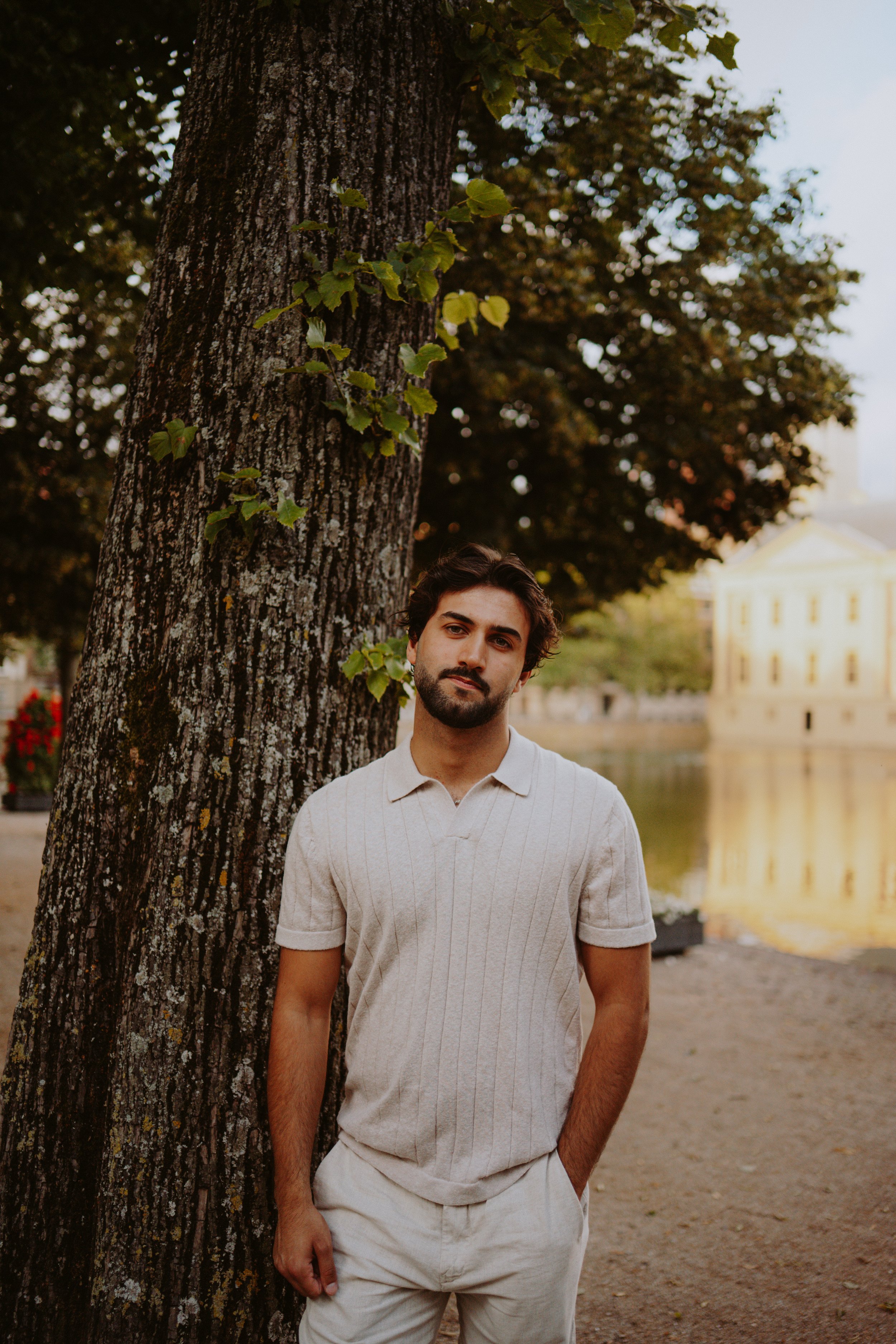 A young man with dark hair and a beard, wearing a cream-colored polo shirt, stands outdoors near a large tree with green leaves. There is a body of water and a beige building in the background.