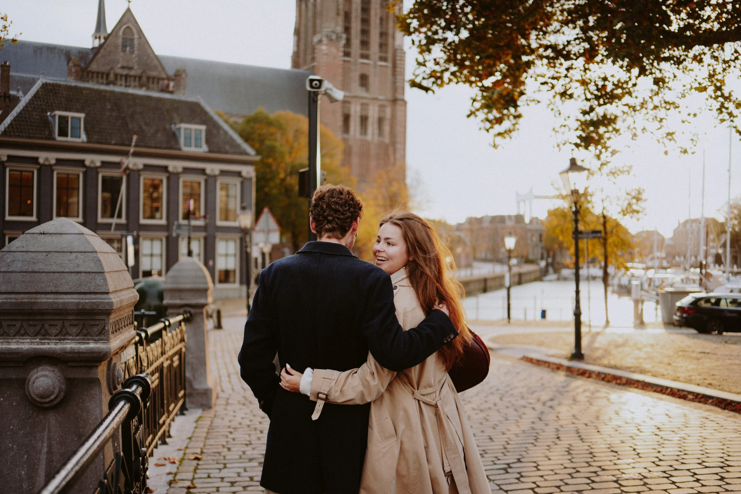 A couple, a man and a woman, embracing and smiling on a bridge in an urban area with historic buildings and fall foliage in the background.