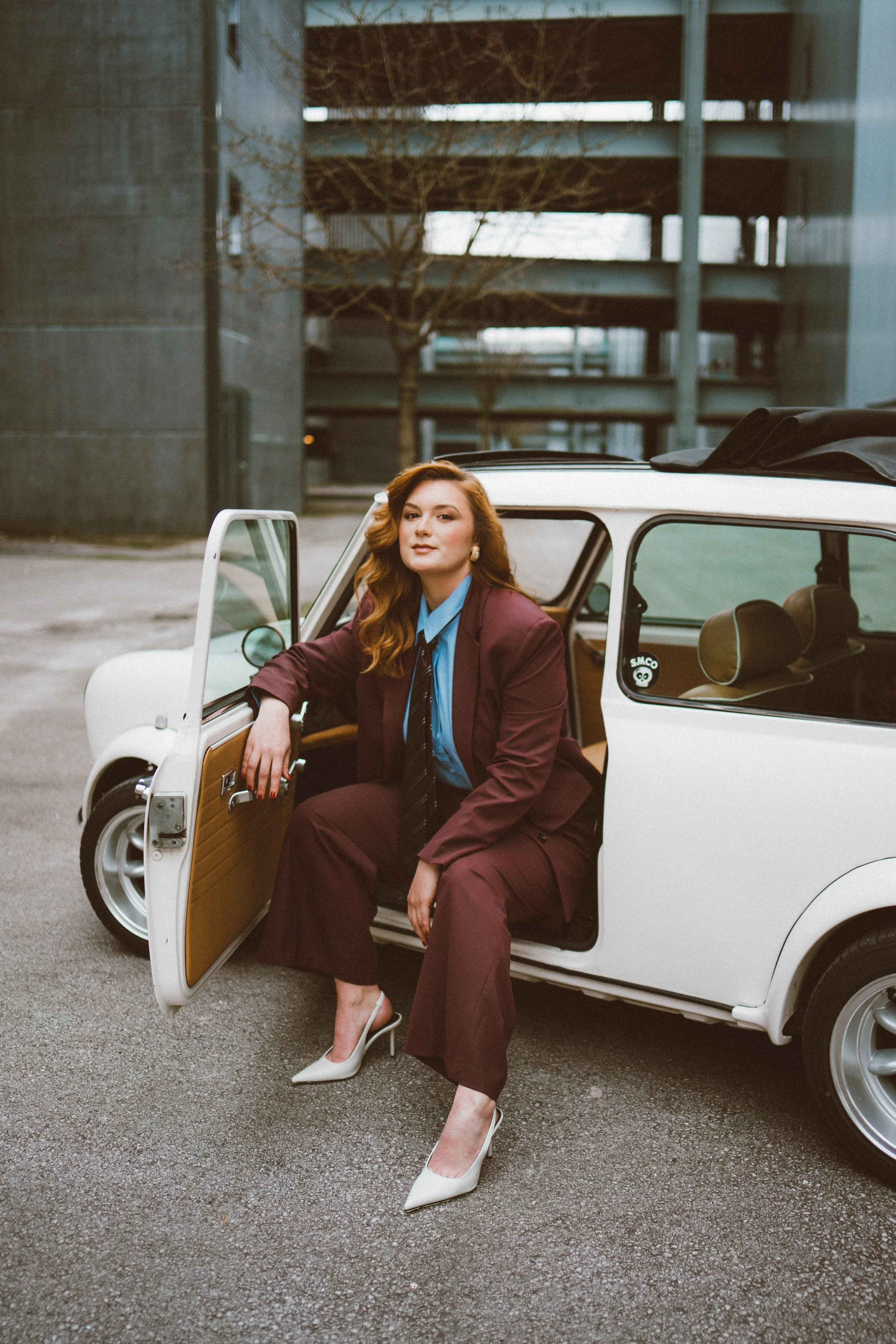 A woman with red hair sitting in a vintage white car with the door open, wearing a burgundy suit and white heels, in an urban setting with modern parking garage in the background.