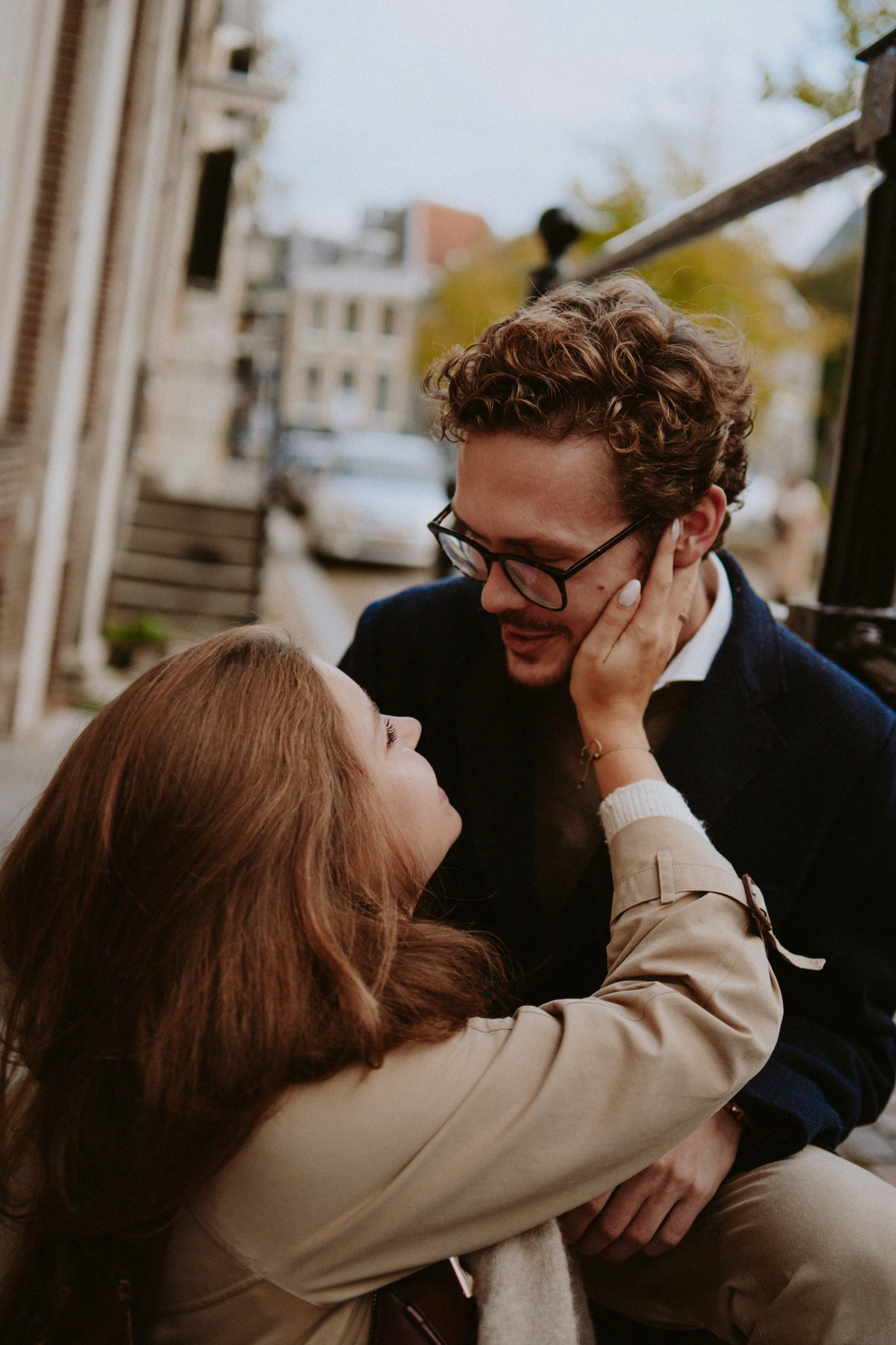 A man with curly hair and glasses is gently touching a woman's face while she is smiling. They are outdoors near a building with a railing in the background.