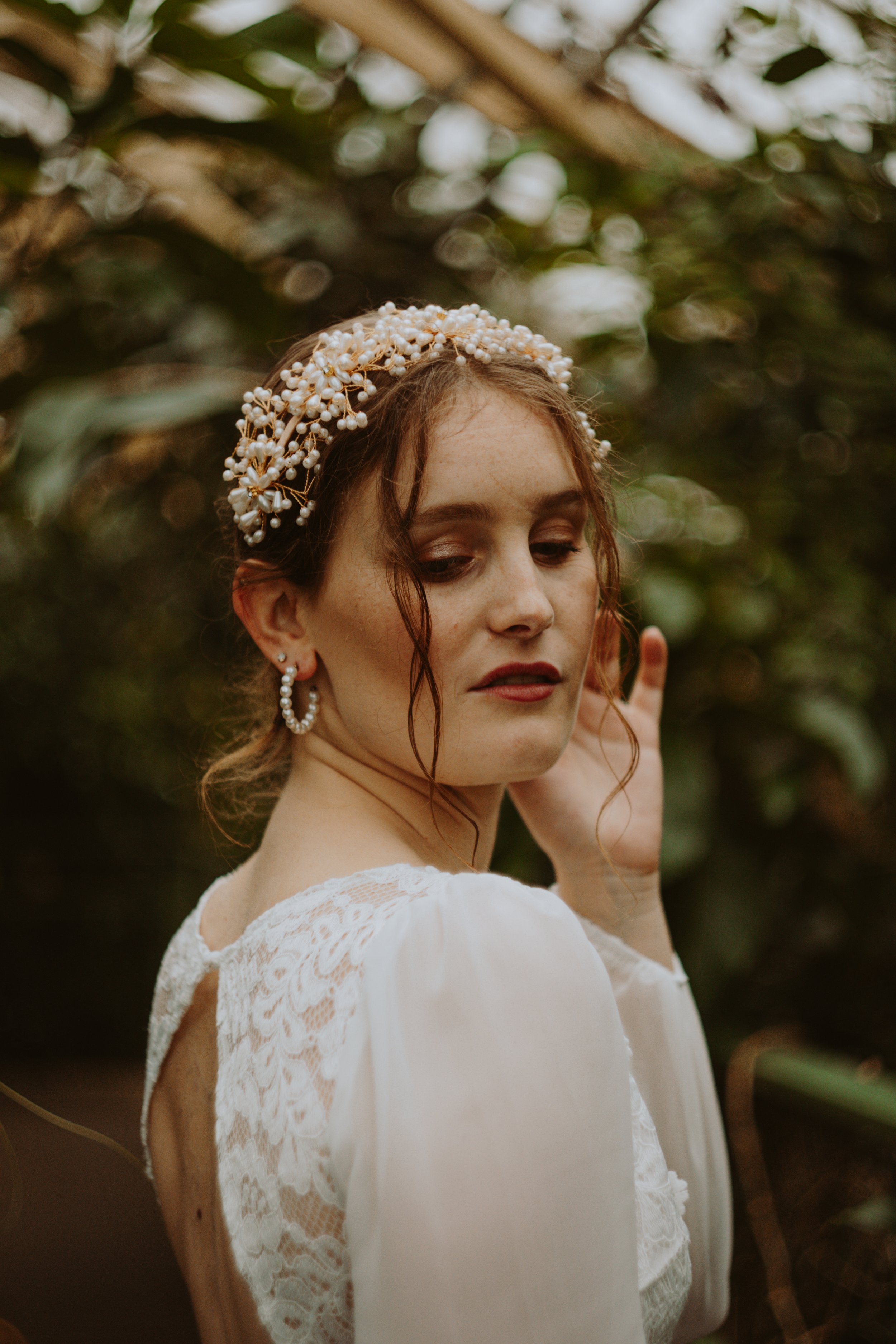 A woman with curly red hair wearing a pearl and floral headpiece and pearl earrings, dressed in a white lace dress, standing outdoors amid green foliage.