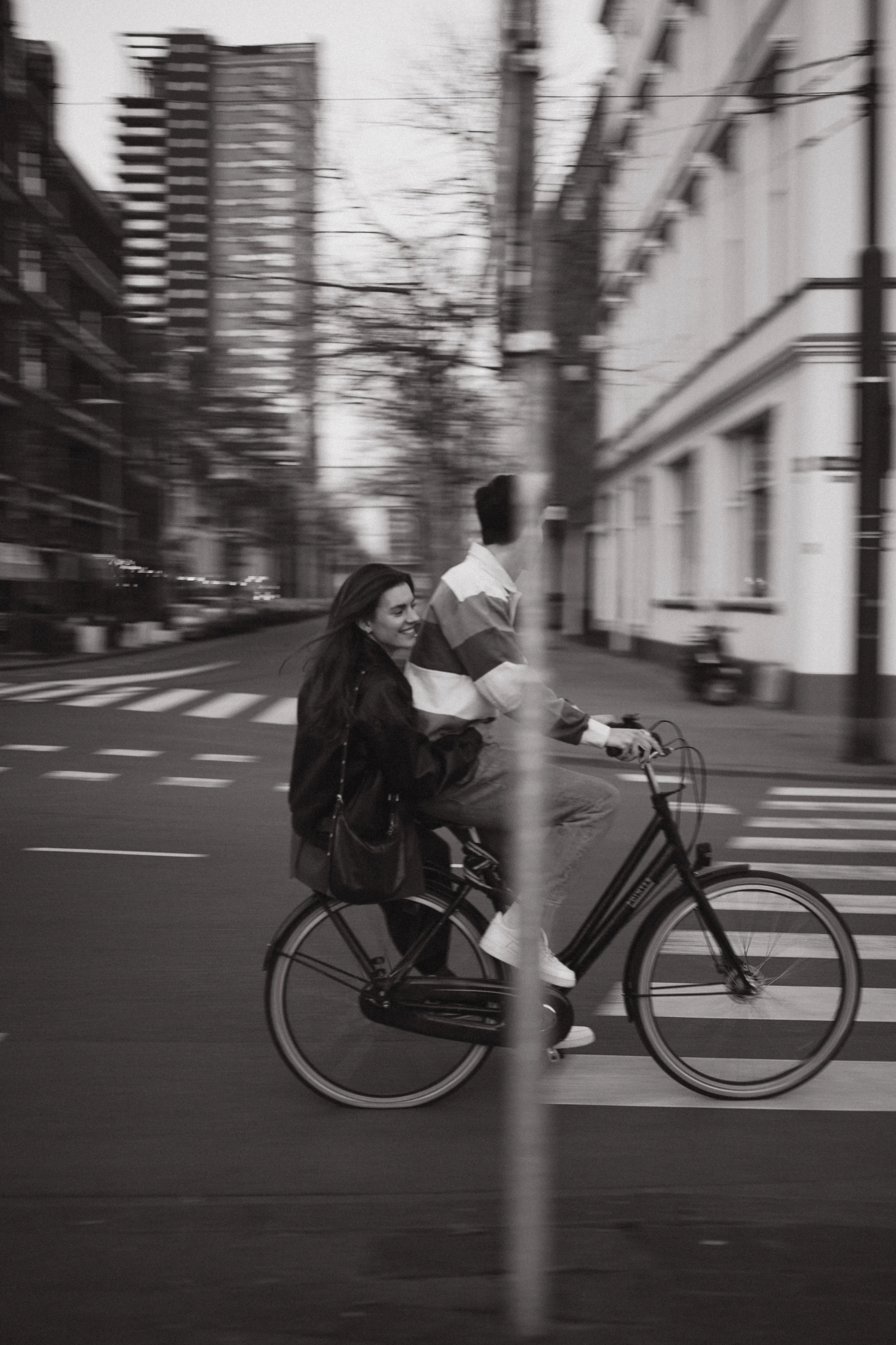 A woman and a man ride a bicycle together on a city street, with tall buildings and leafless trees in the background, captured in black and white, motion-blurred.