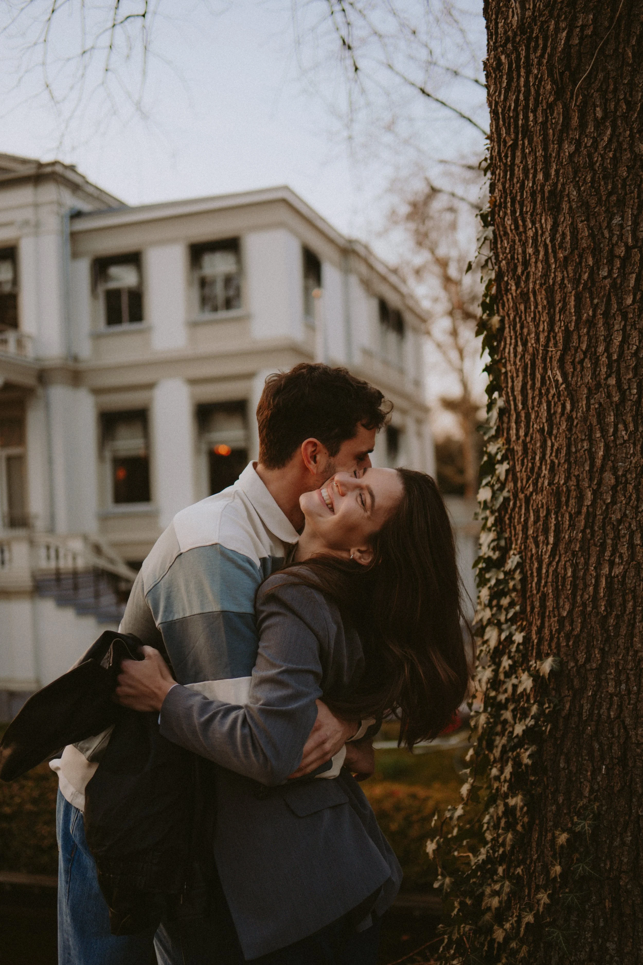 A couple is embracing and smiling near a large tree with ivy, with a white house and trees in the background during dusk.