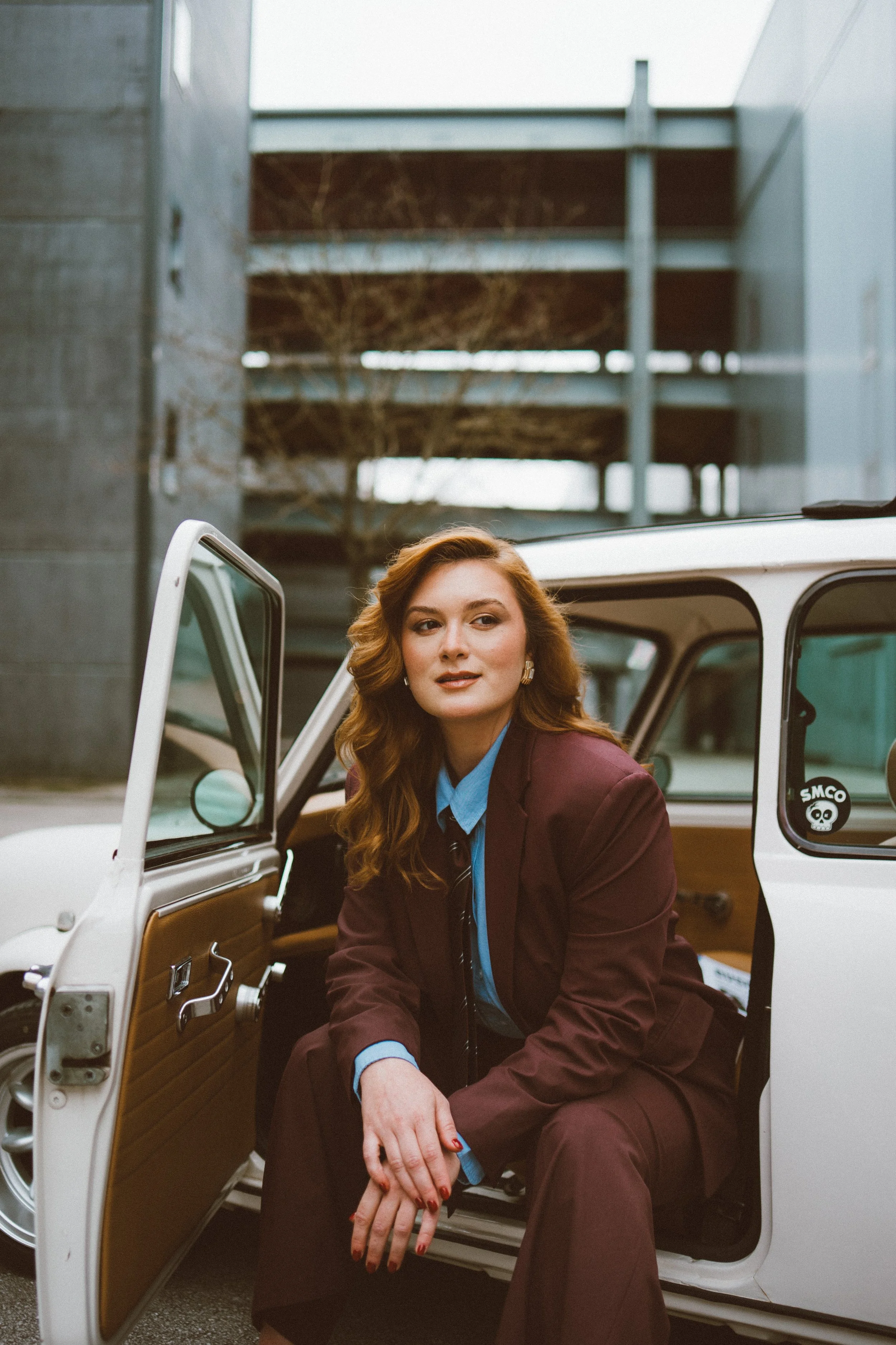 Photo of a business woman with red hair wearing a maroon suit and blue shirt sitting in the open door of a white vintage car with wooden interior. The background features a multi-story parking garage and leafless trees.
