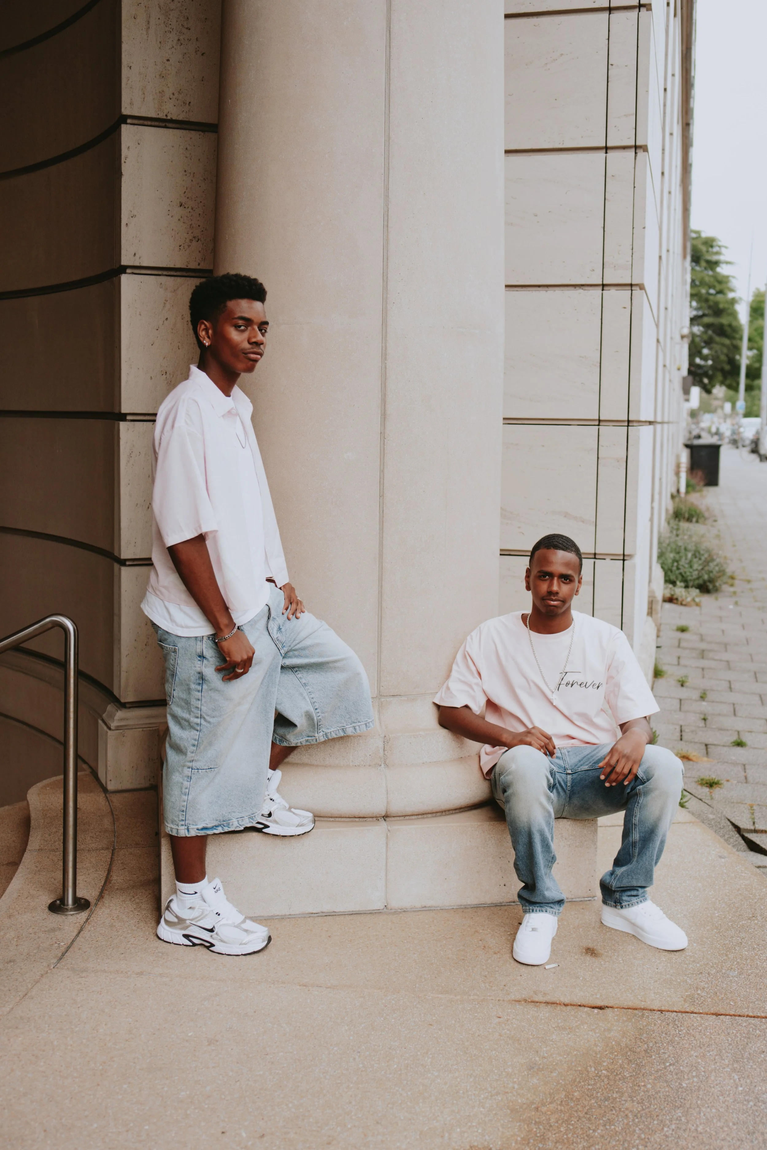 Two young men are posing outside near a building with beige stone walls. One is leaning against the wall, wearing a white shirt, oversized denim shorts, and Nike sneakers. The other is sitting on a ledge, wearing a pink t-shirt with 'Forever' written