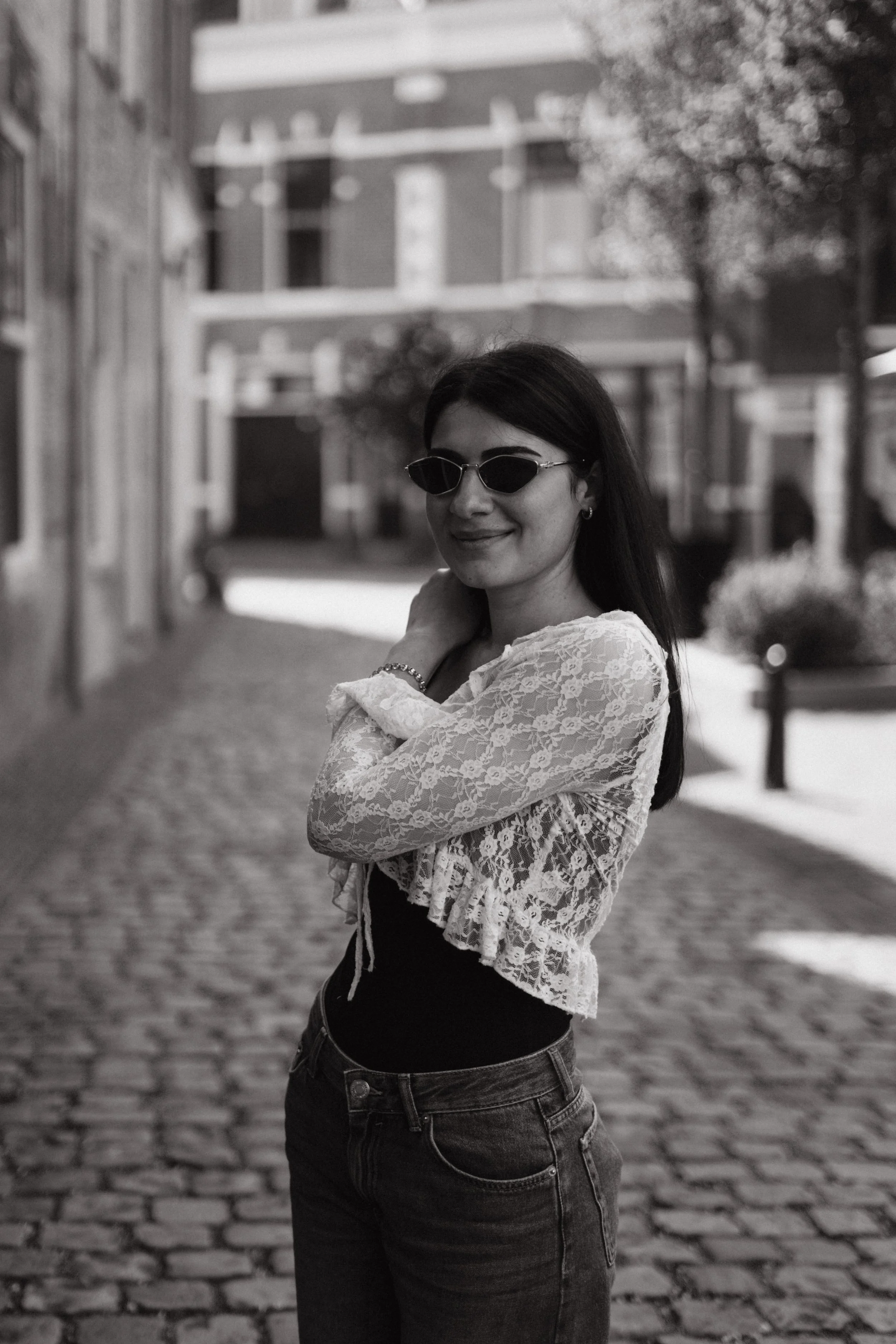 A woman with dark hair wearing sunglasses, a lace top, a black tank top, and jeans standing on a cobblestone street with blurred buildings and trees in the background.