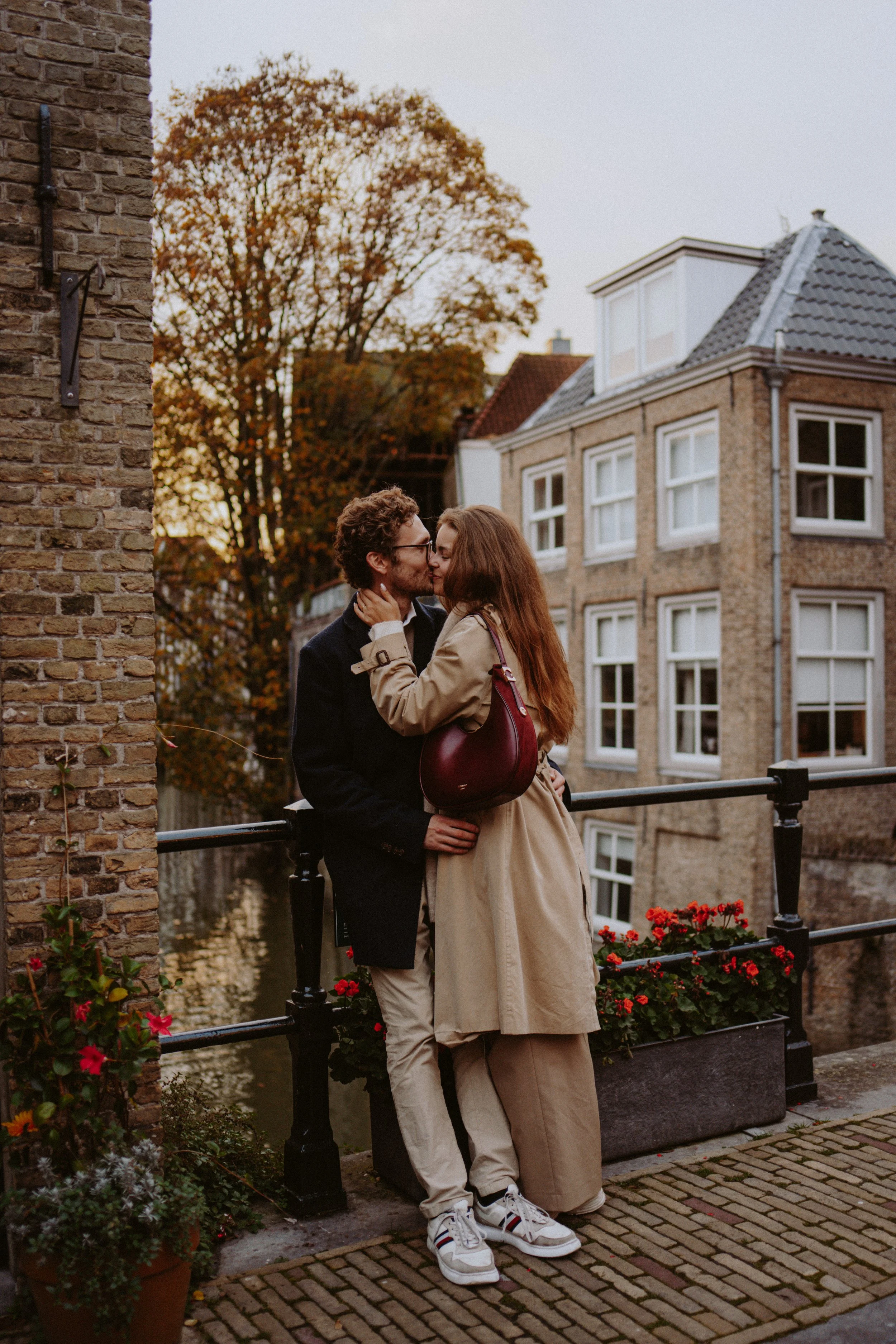 A couple sharing a kiss on a bridge over a canal in an urban area during autumn, with brick buildings and a large tree with colored leaves in the background.