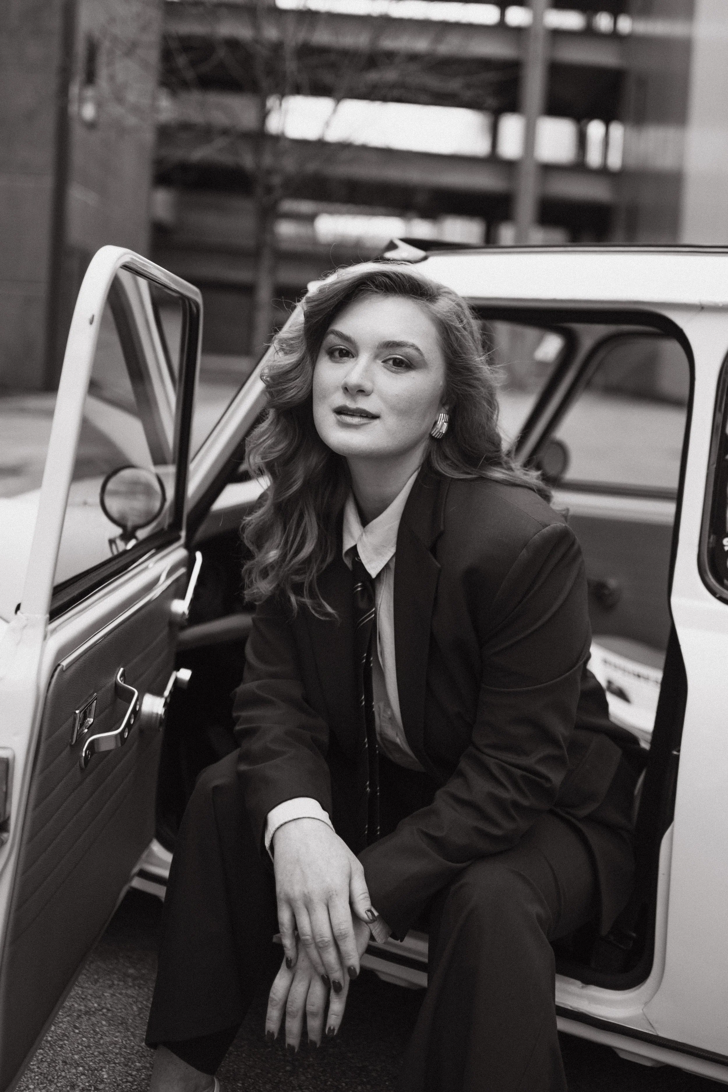 Black and white portrait of a woman with wavy hair, wearing a suit and tie, sitting in a vintage car with the door open. 