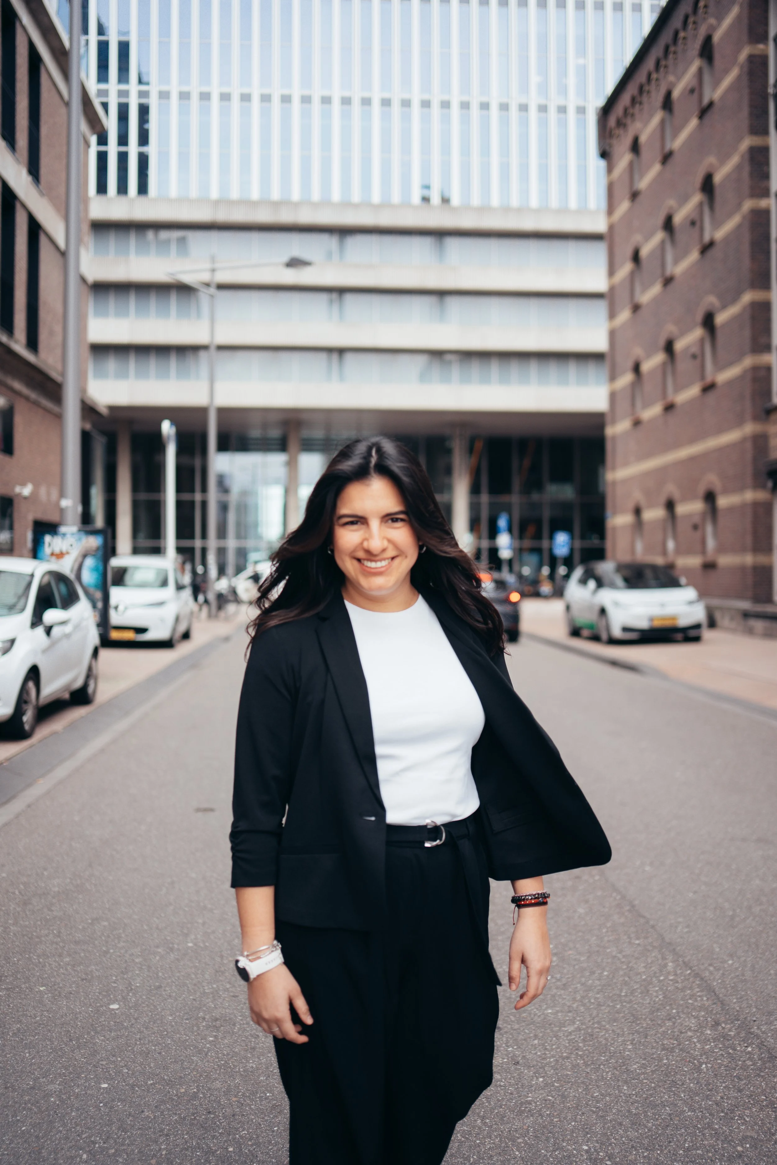 A woman smiling and walking on a city street, wearing a black blazer, white shirt, and black pants with architectural buildings and parked cars in the background.