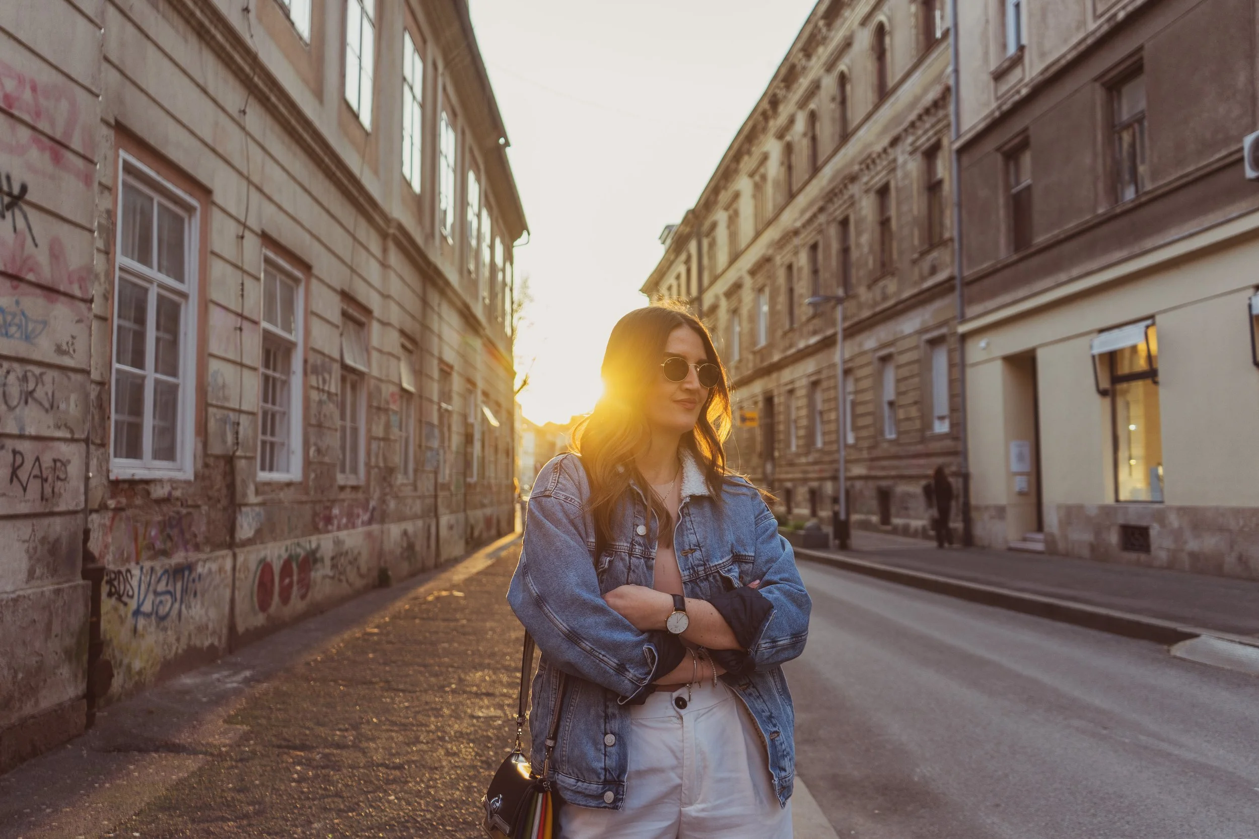 A woman with long dark hair wearing sunglasses, a denim jacket, and white pants, standing with arms crossed on a city street during sunset with old buildings and graffiti on the wall behind her.