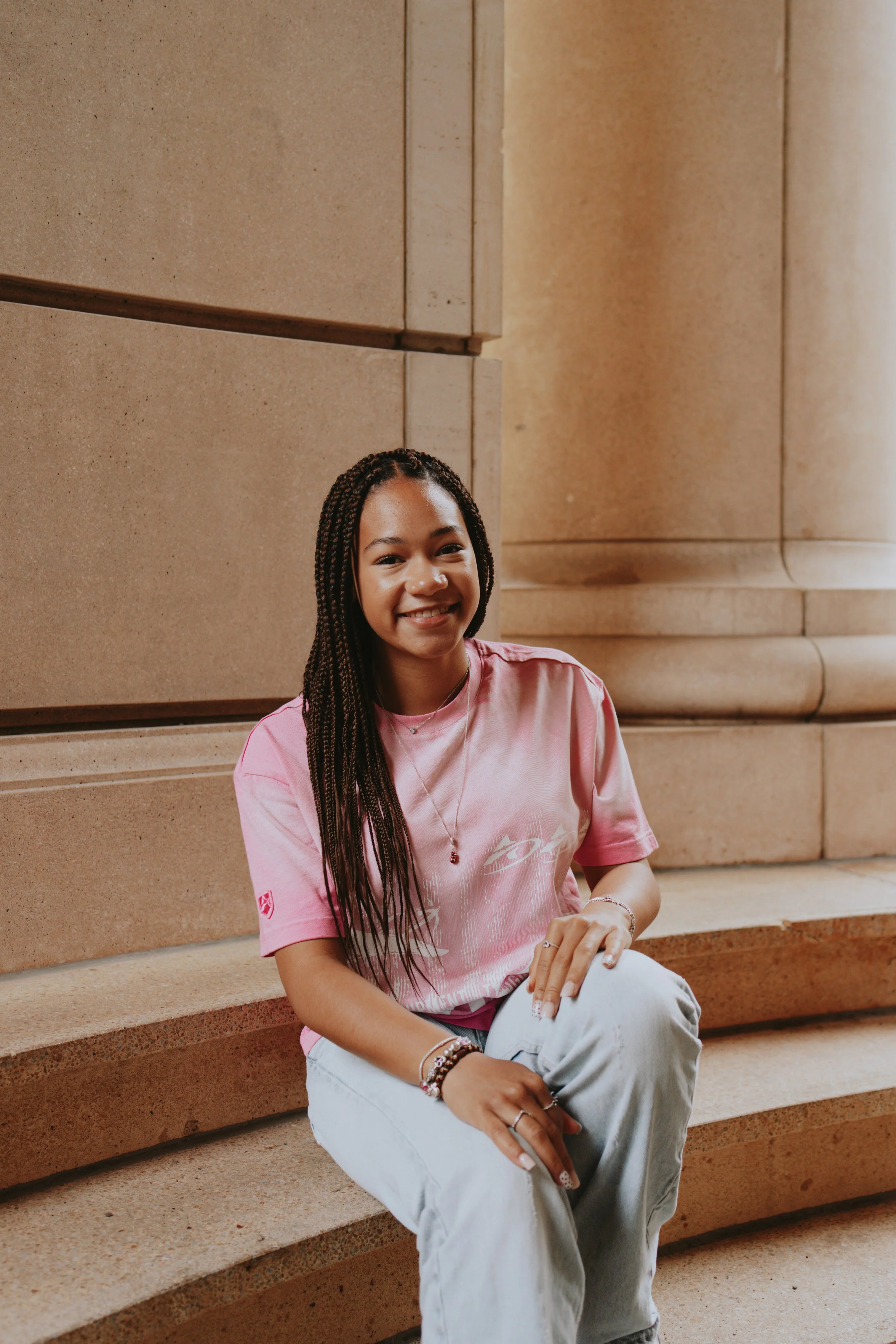 A young woman with long braided hair and a pink T-shirt sitting on stone steps outdoors, smiling at the camera.