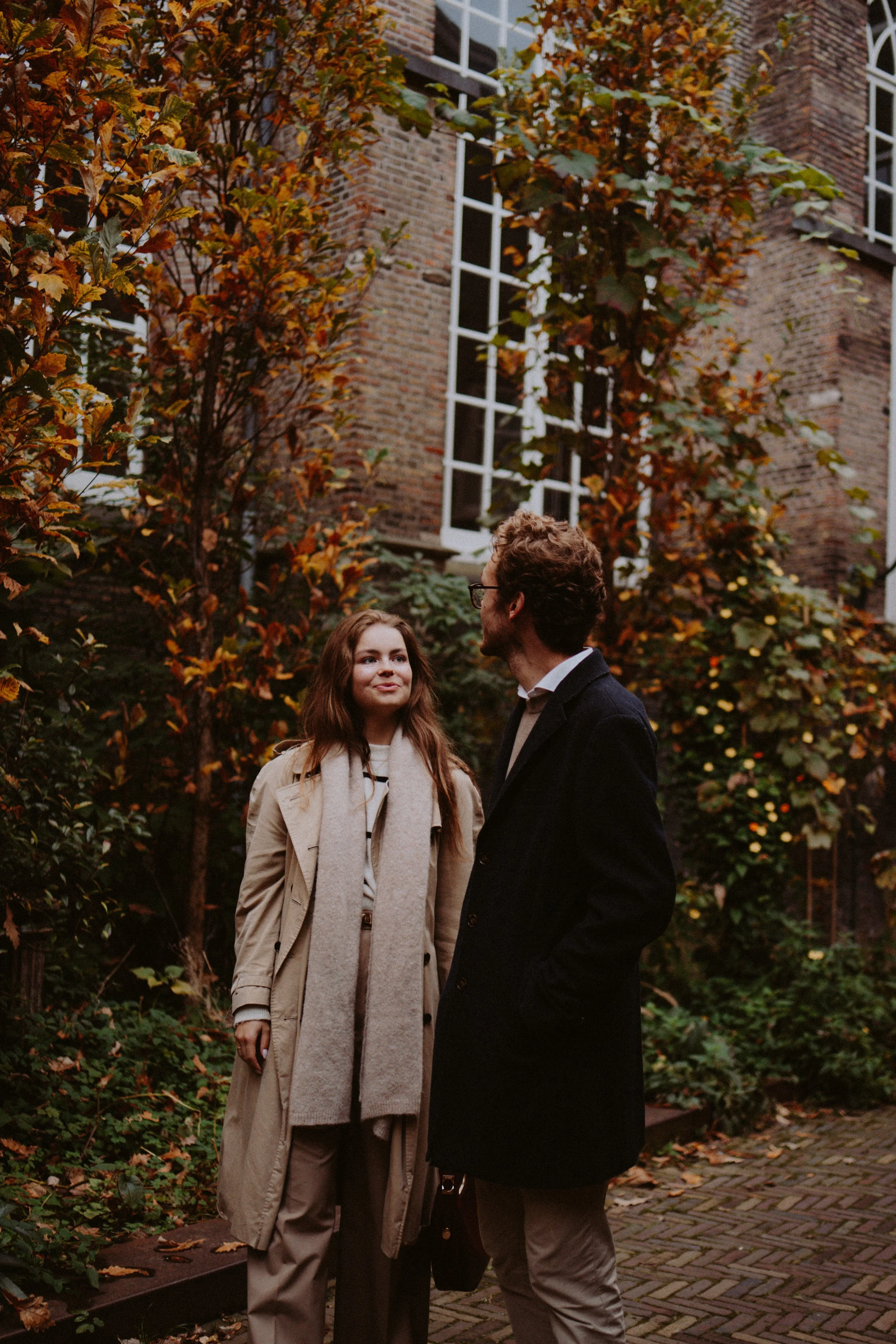 A man and woman are standing outdoors among autumn foliage in front of a brick building with large windows, engaging in conversation.