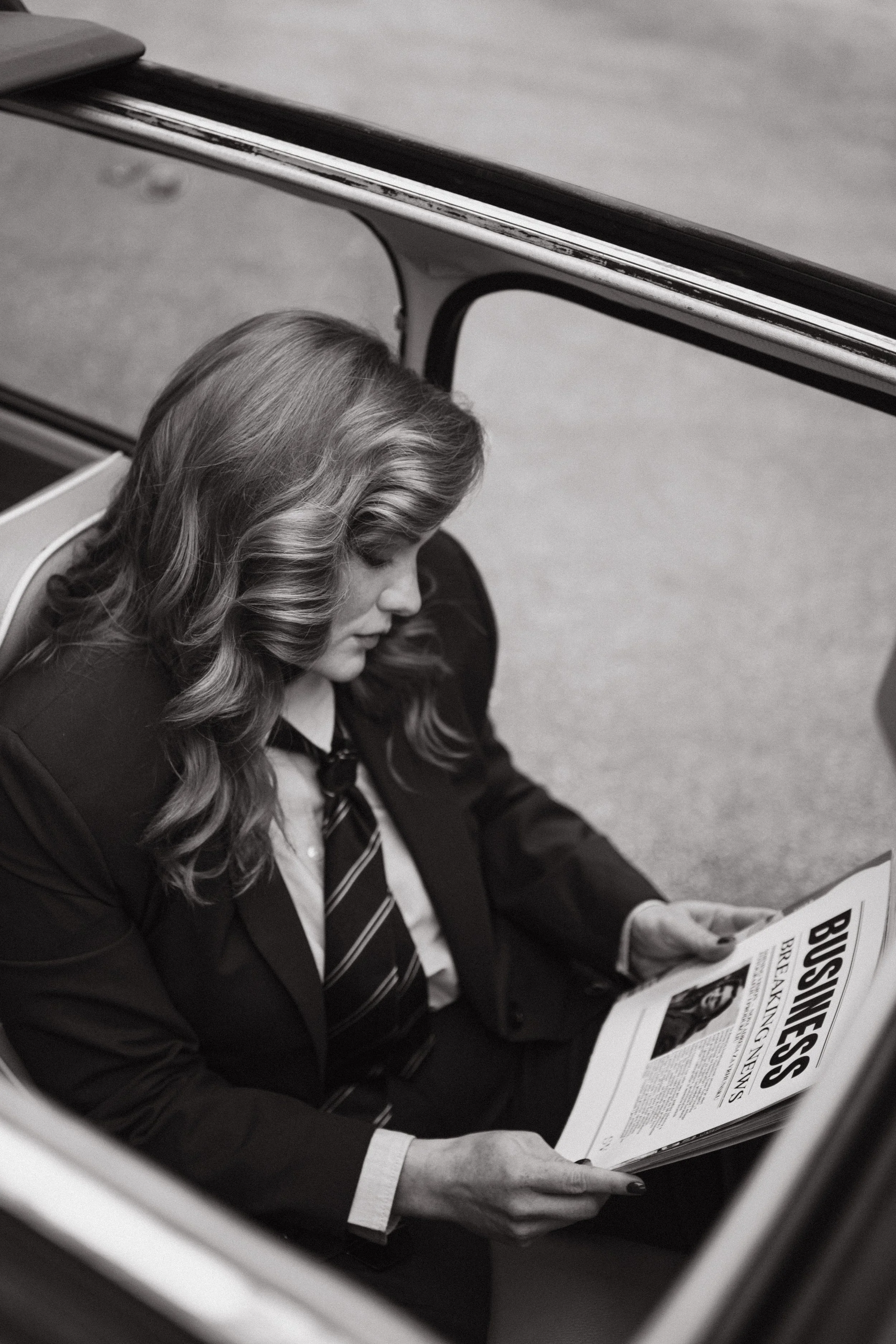 Black and white photo of a woman in formal attire sitting under a desk, reading a newspaper titled 'Business'.